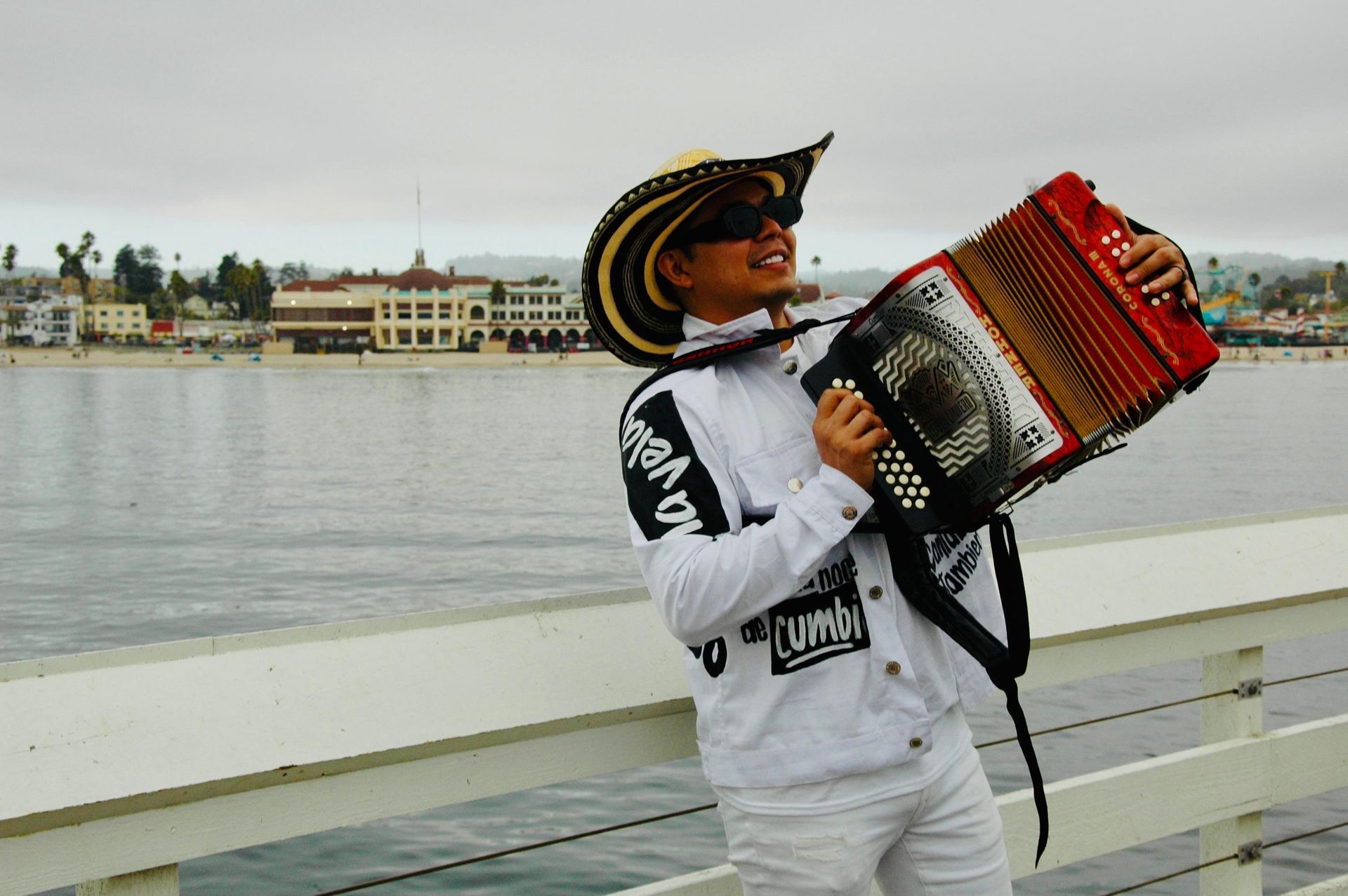 Un hombre toca el acordeón en el muelle, sonriendo, vistiendo sombrero y chaqueta blanca, con el océano y edificios de fondo.