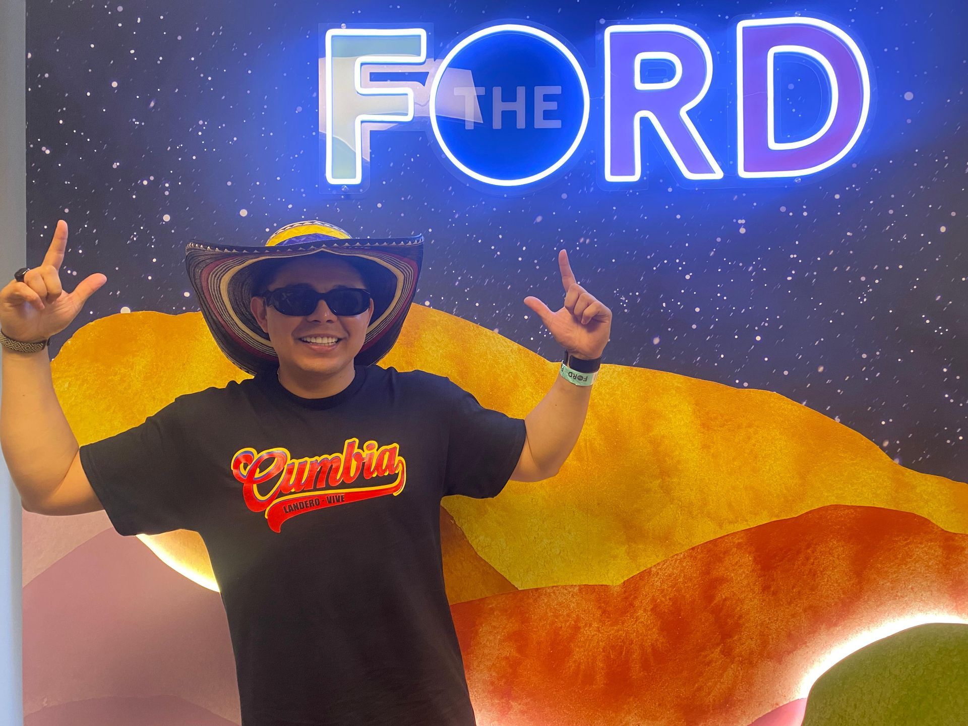 Persona con sombrero y gafas de sol posa frente a un cartel de