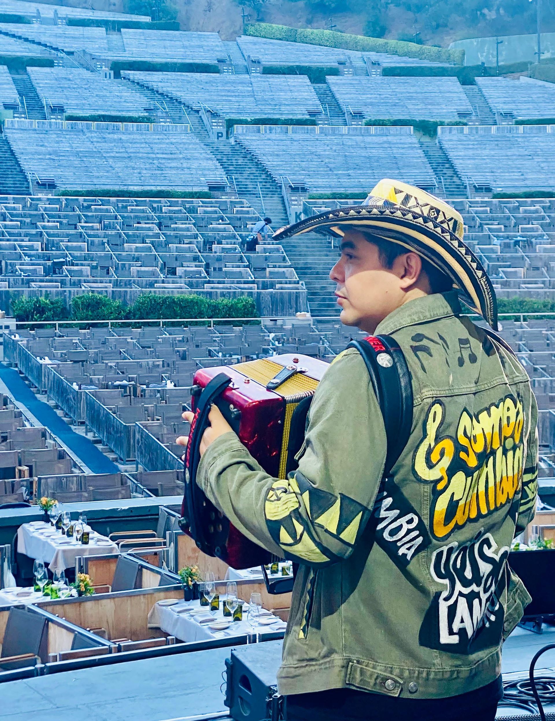 Un hombre con sombrero de vaquero toca el acordeón al aire libre; un anfiteatro al fondo.