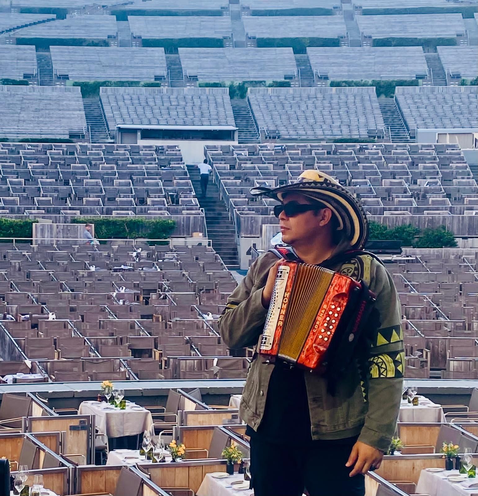 Un hombre con acordeón y sombrero se encuentra en un estadio, sosteniendo el instrumento con una audiencia en el fondo.