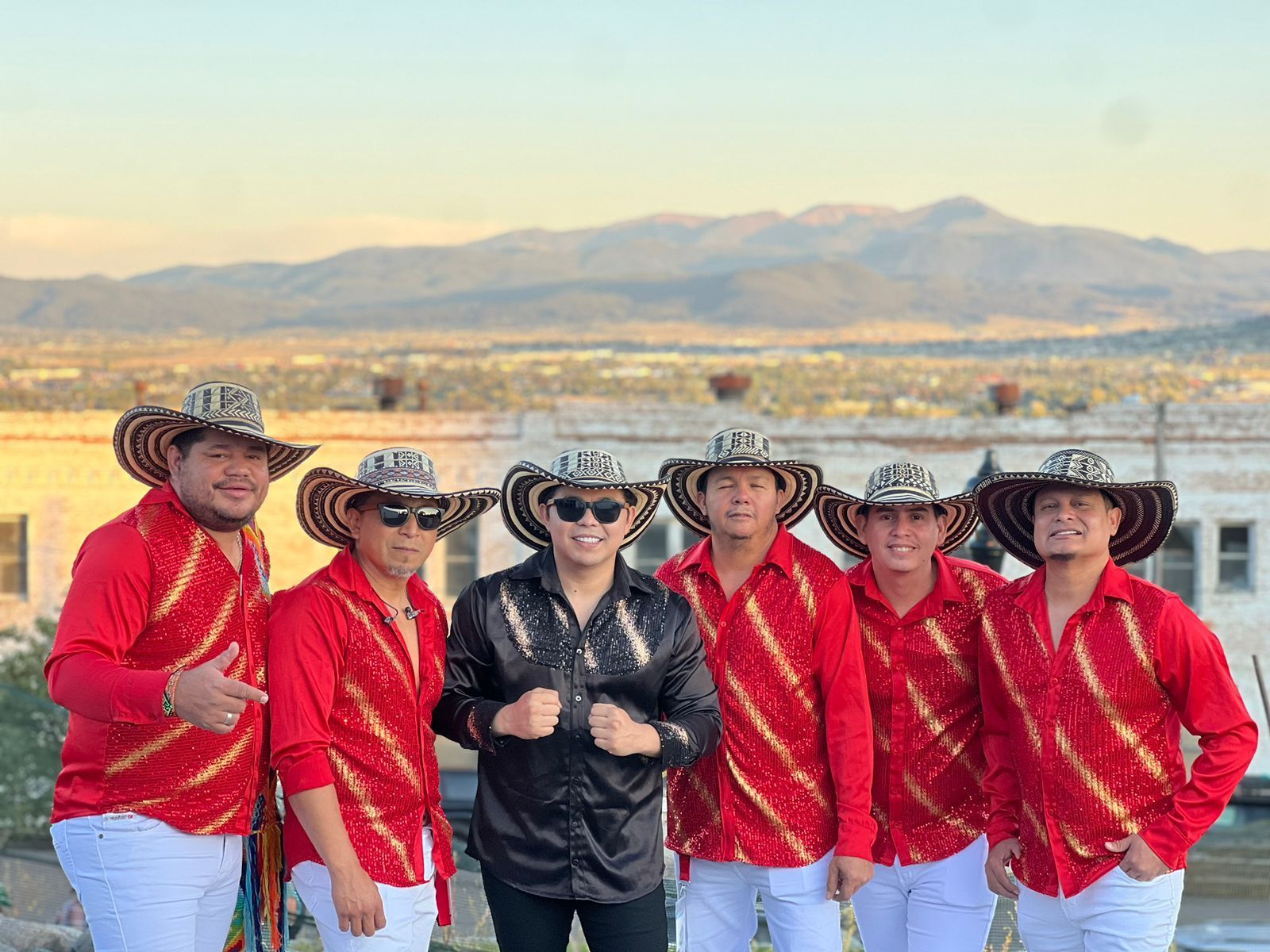 Seis músicos con camisetas y sombreros rojos posan al aire libre con un fondo de montañas.