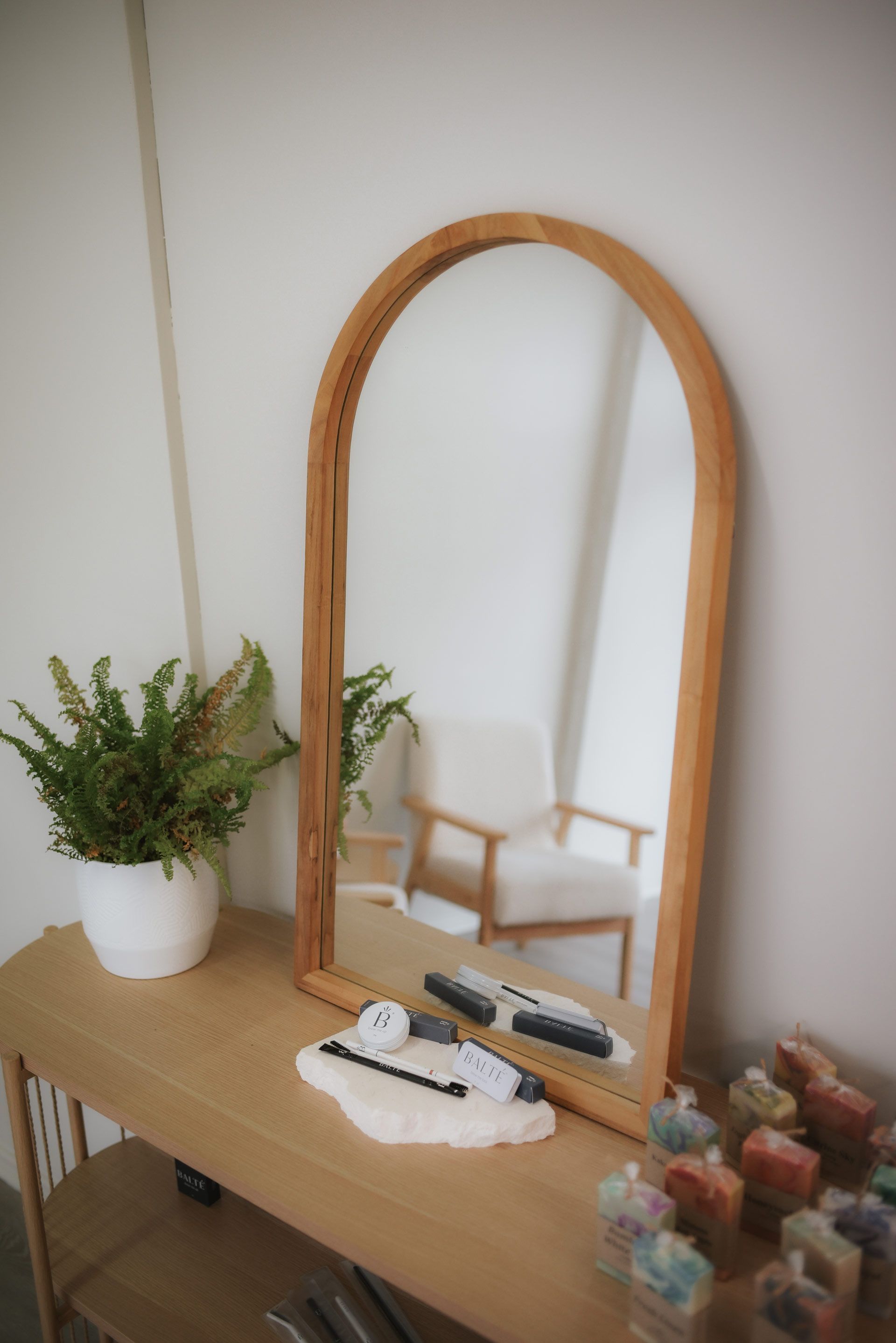Wooden arch mirror on a table with plants and decor, reflecting a chair and bright room.