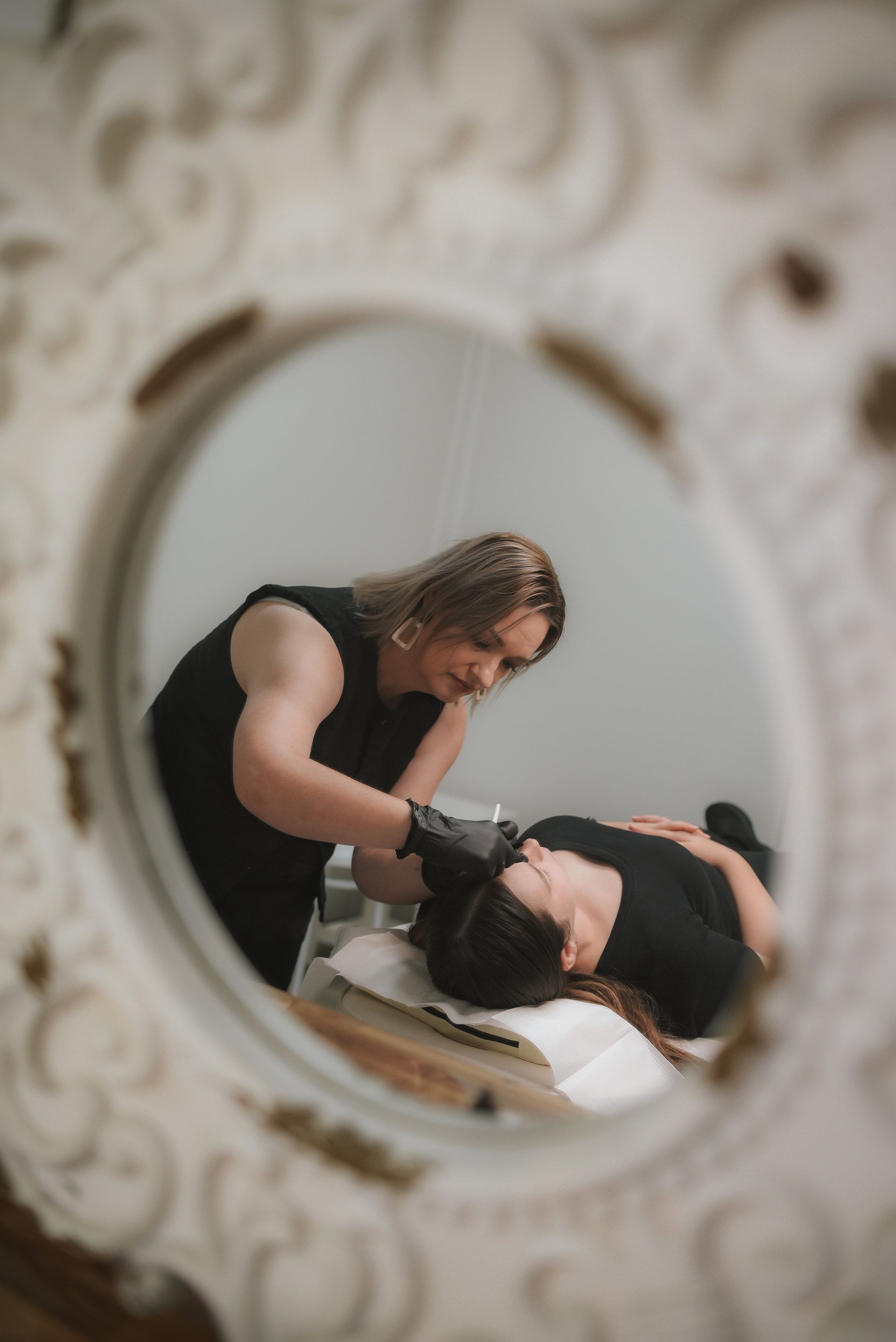 A person working on a client's face in a salon, reflected in a decorative mirror.