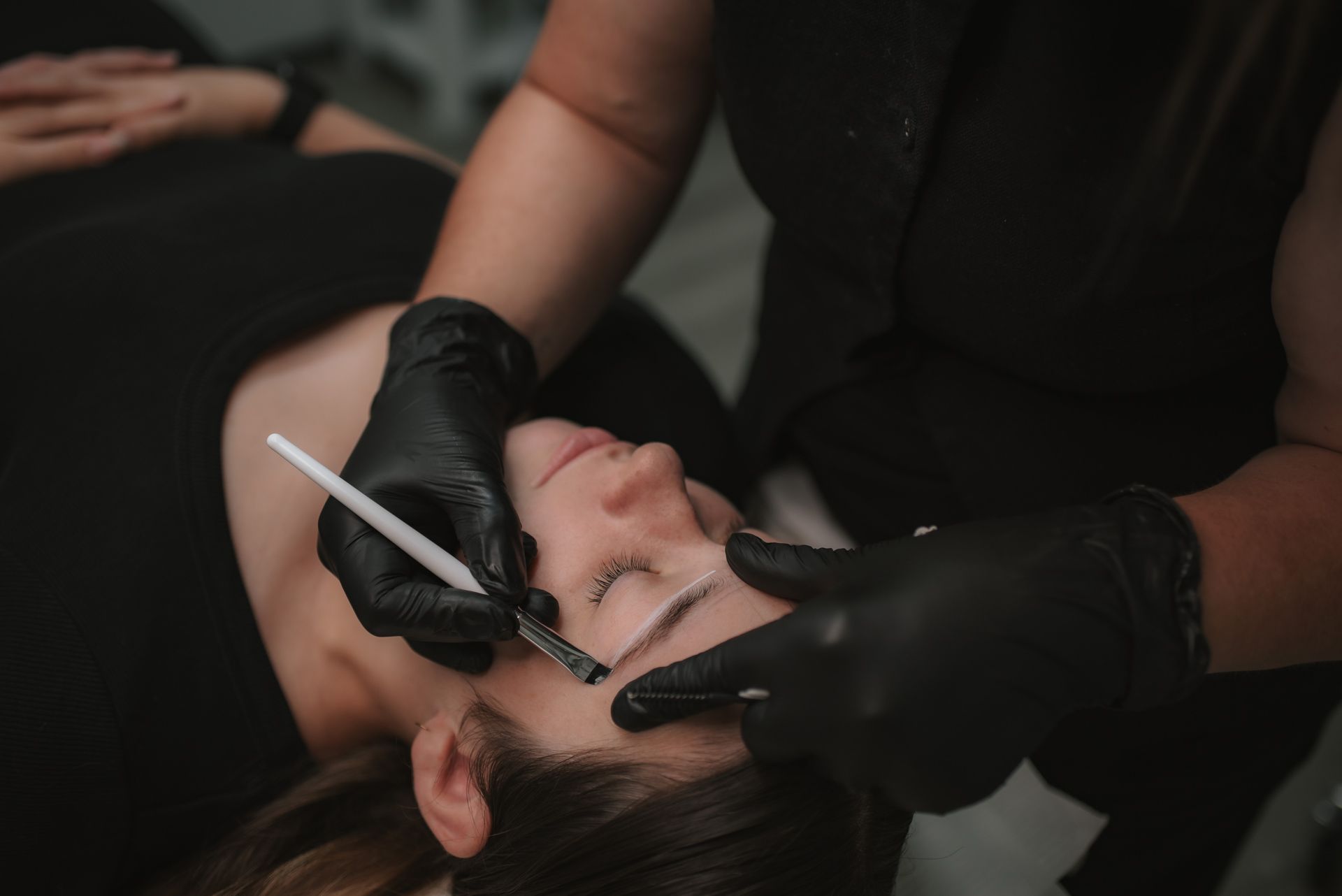 A woman getting her eyebrows shaped by a professional wearing black gloves; indoor setting.