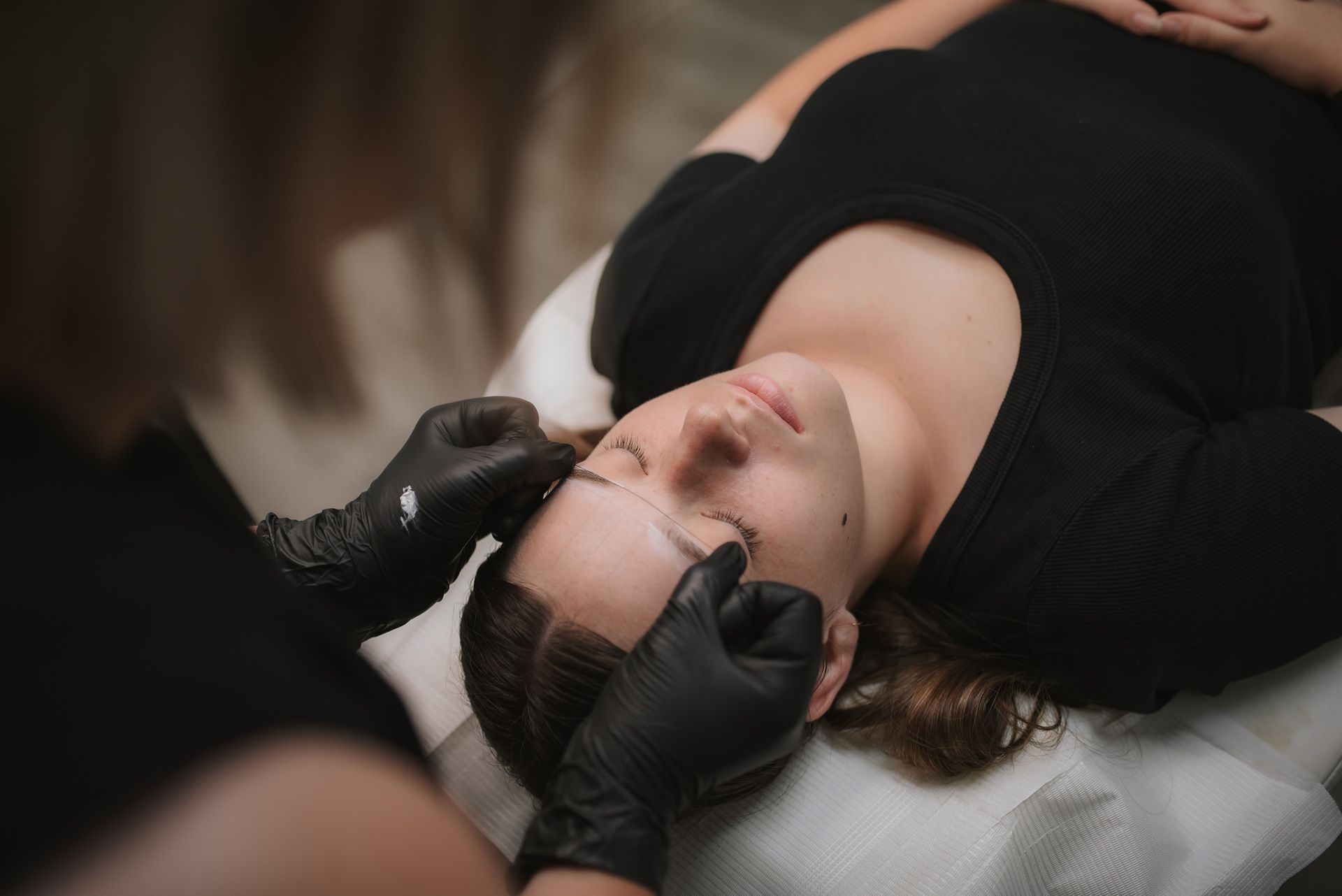 A woman lying down while a person with black gloves shapes her eyebrows with a string.