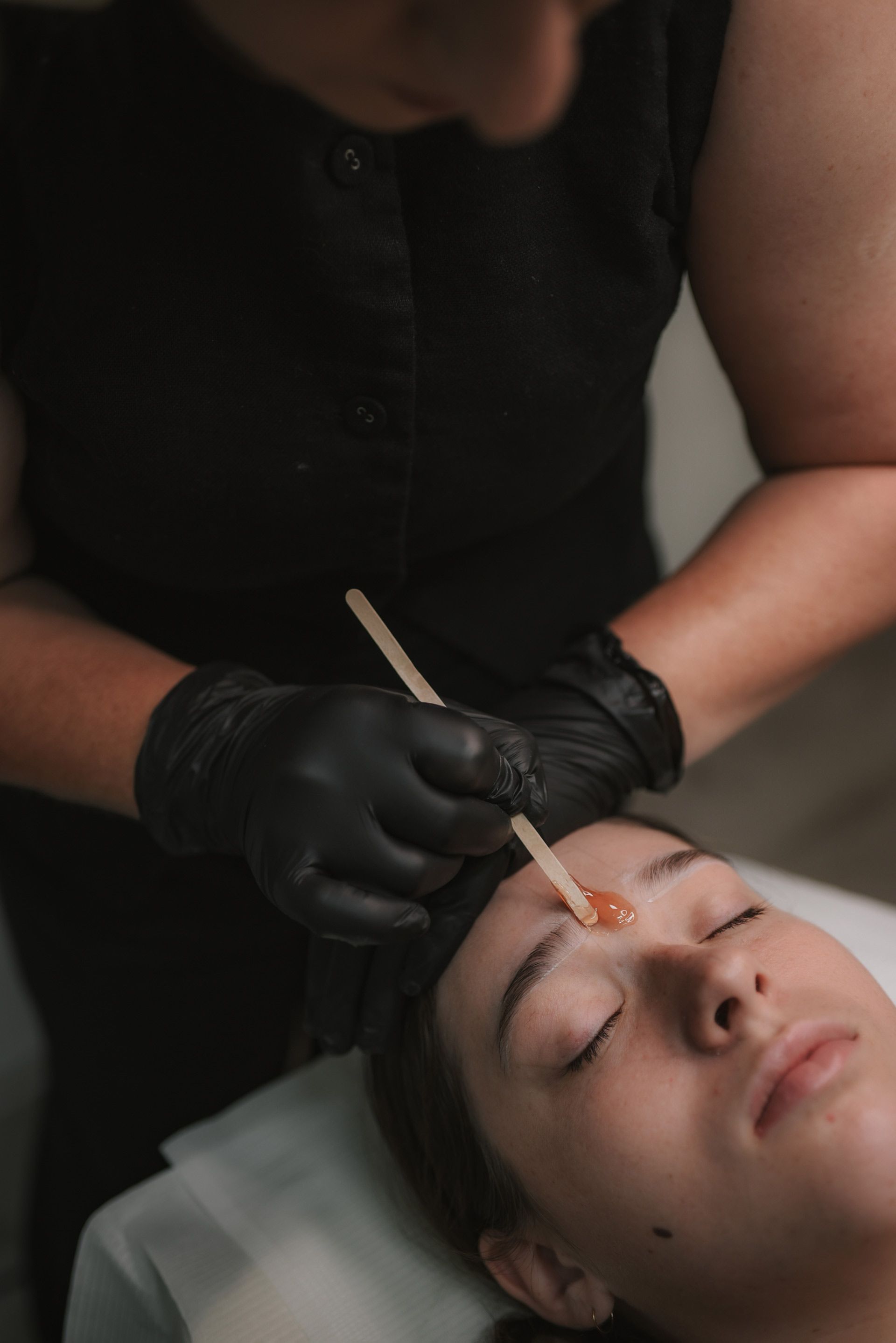 Woman receiving eyebrow waxing in a salon; aesthetician wears black gloves, holding a wax applicator.