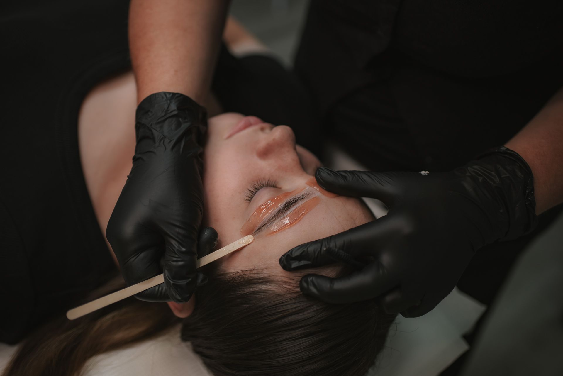 Person having eyebrow waxing; hands in black gloves applying wax.