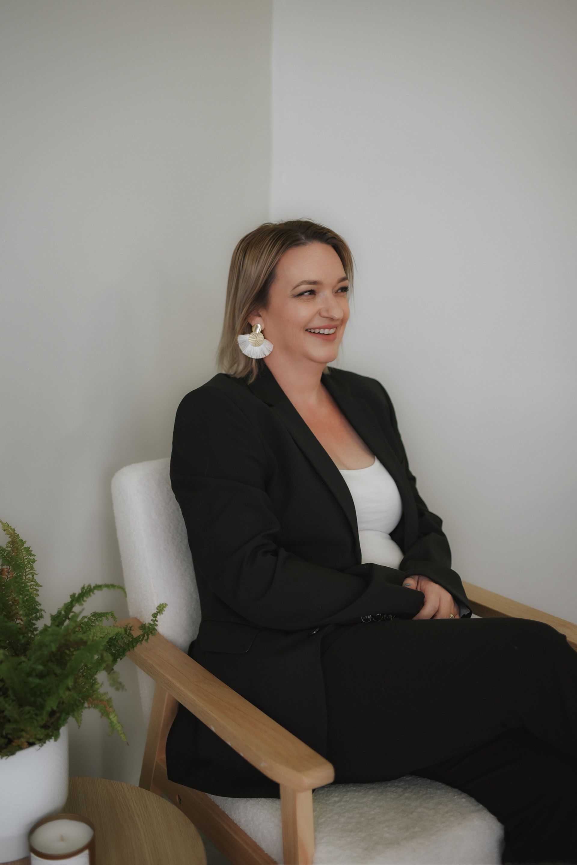 Woman in black suit sitting in chair, smiling in corner of white room.