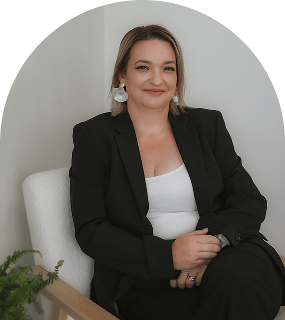 Woman in black suit sitting in a white chair, smiling, with a neutral background.