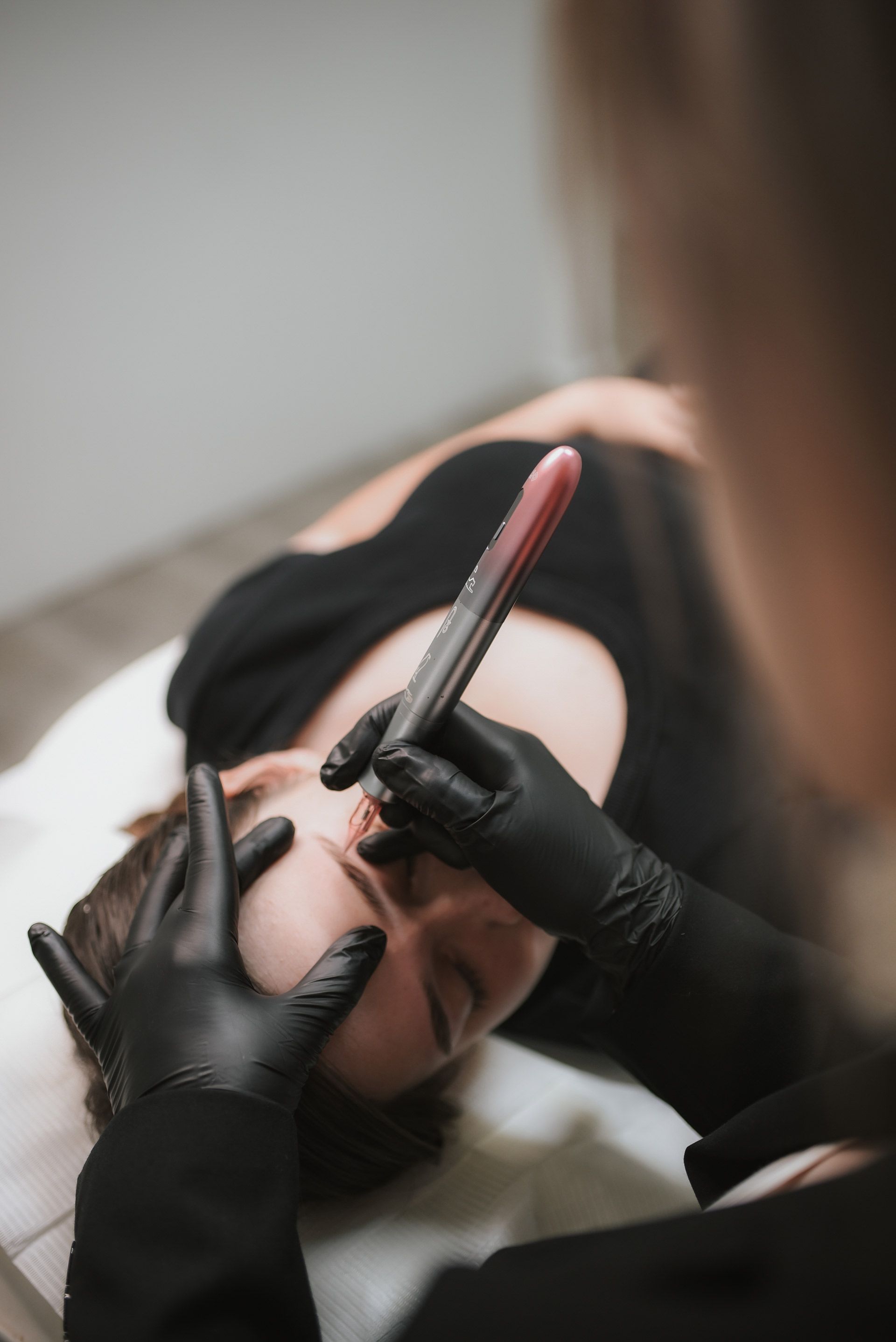 Woman receiving eyebrow microblading; artist in black gloves holding tool over her brow in a salon.
