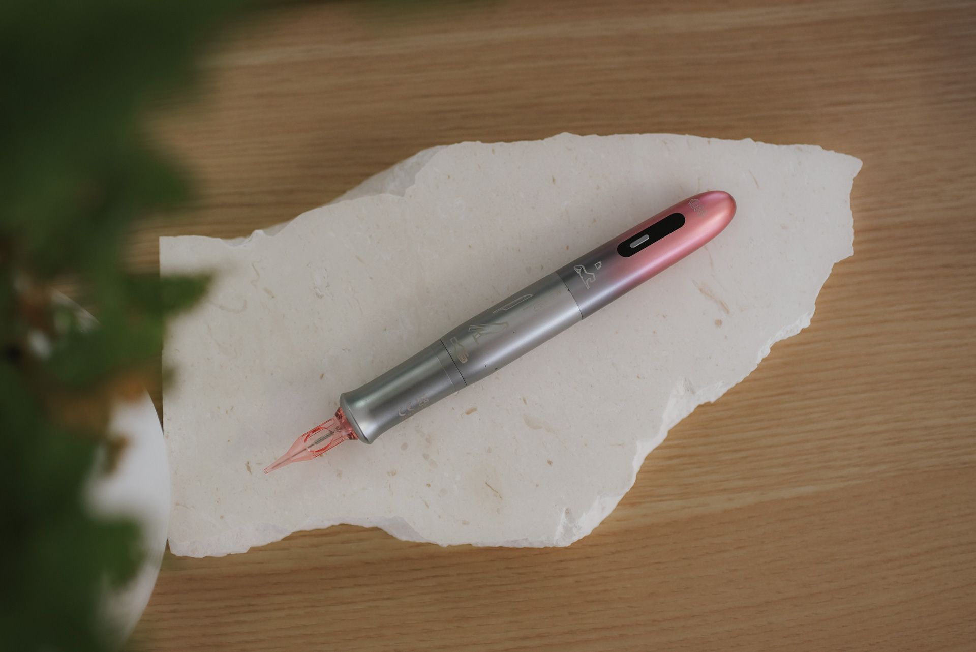 Pink and silver pen on a white, irregularly shaped stone, resting on a wooden surface, with greenery on the left.