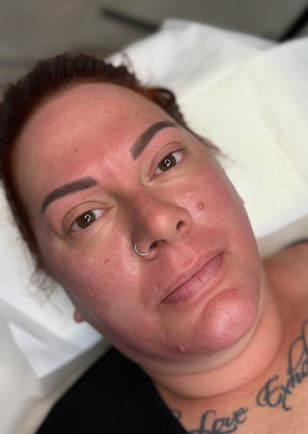 Woman with red skin, freshly done eyebrow tattoo, nose ring, and chest tattoos, laying down.