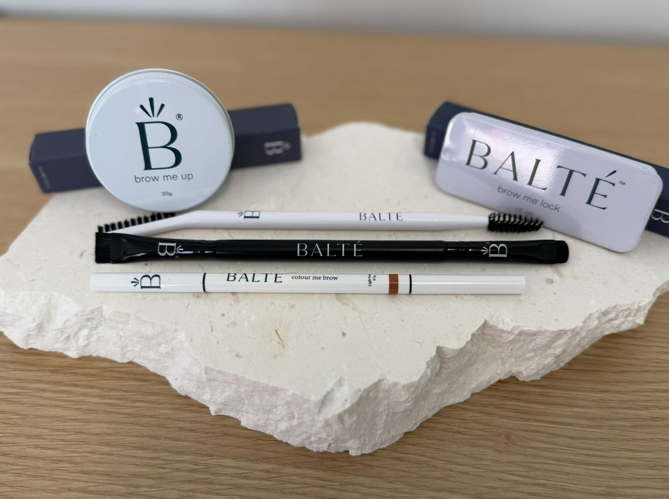 Beauty products set on a white stone: eyebrow gel, pencil, and brush. The products are in white and black packaging.