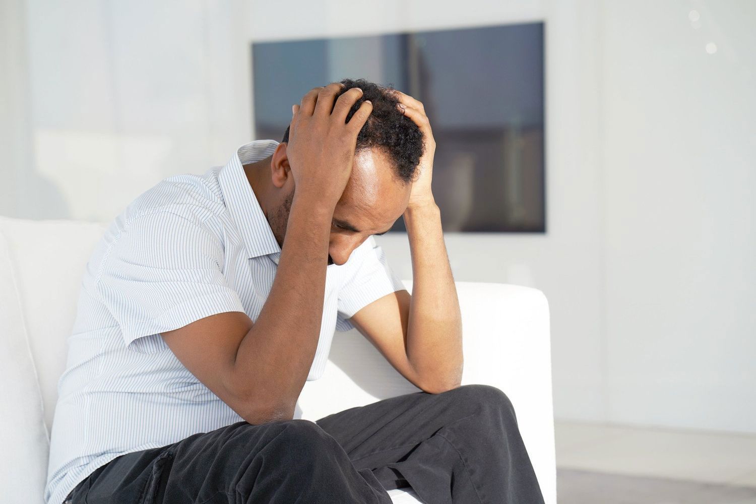 A man with dark skin sits on a white couch, holding his head in his hands with a distressed expression.