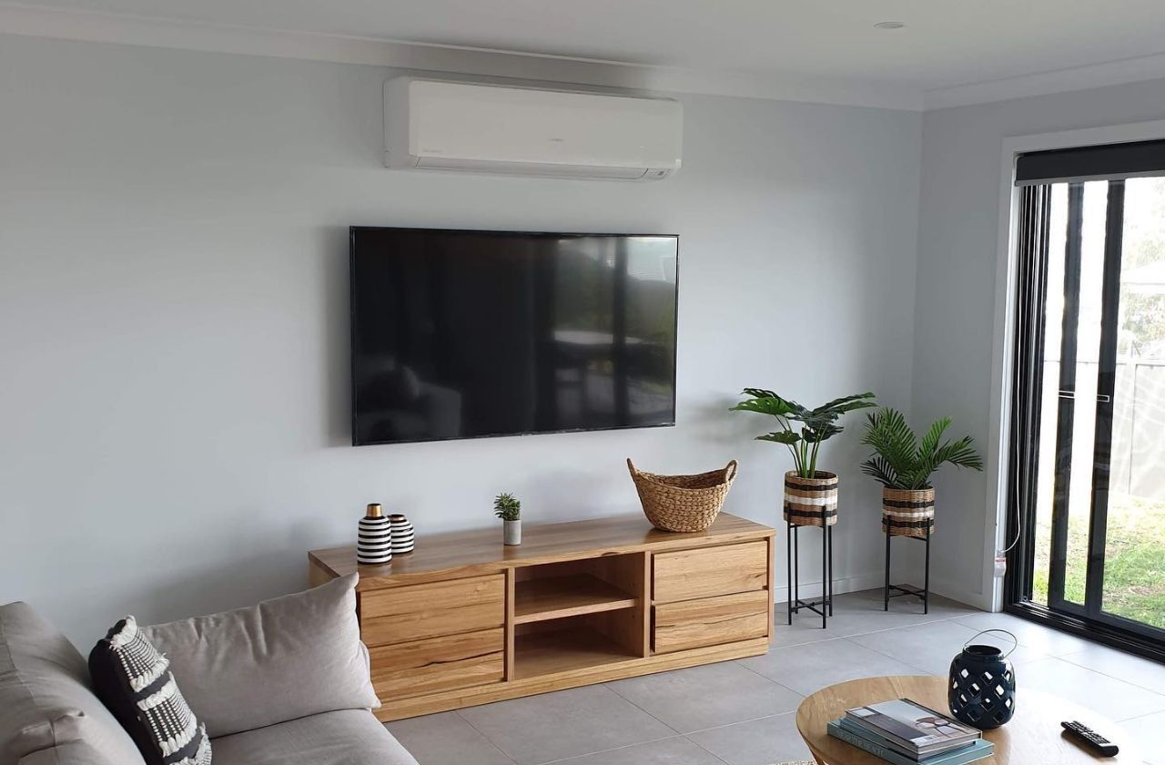 Living room with mounted TV, wooden cabinet, air conditioner, plants, and sliding glass door. — Bay & Basin Air Conditioning & Refrigeration Pty Ltd in St Georges Basin, NSW