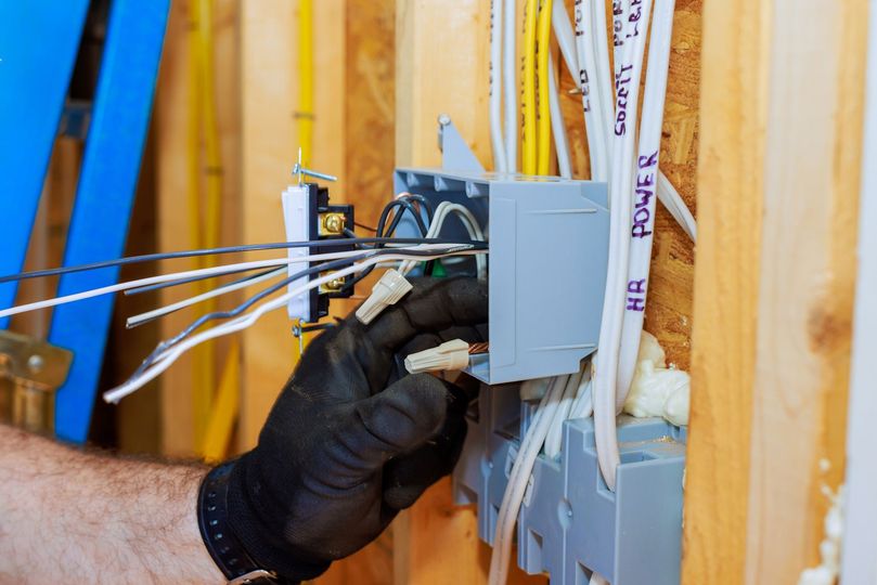 Electrician connecting wires in a wall-mounted electrical box. Hands in black gloves; wires and box are gray, white, and yellow.