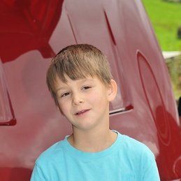 Un jeune garçon en chemise bleue est assis devant une voiture rouge.