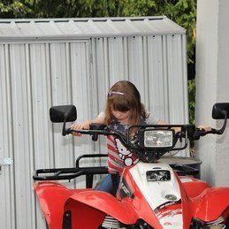Une petite fille conduit un VTT rouge devant un hangar blanc.
