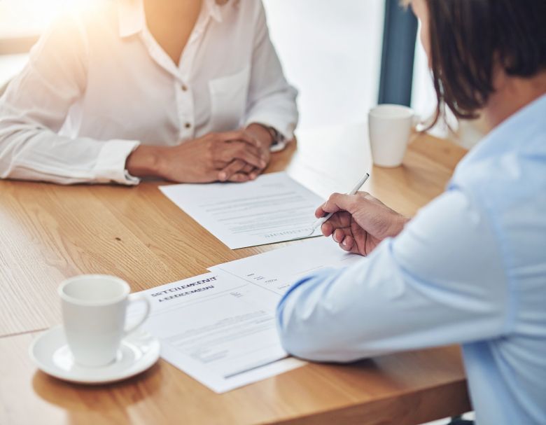Two people seated at a wooden table, signing documents. Cups of coffee are on the table.