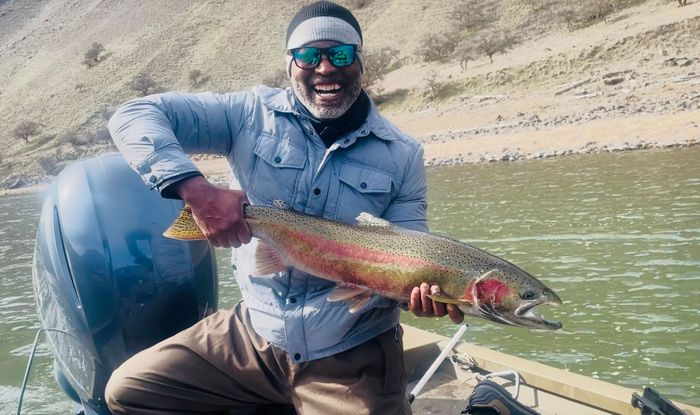 Man on boat holding a large rainbow trout. River in background. He's smiling.