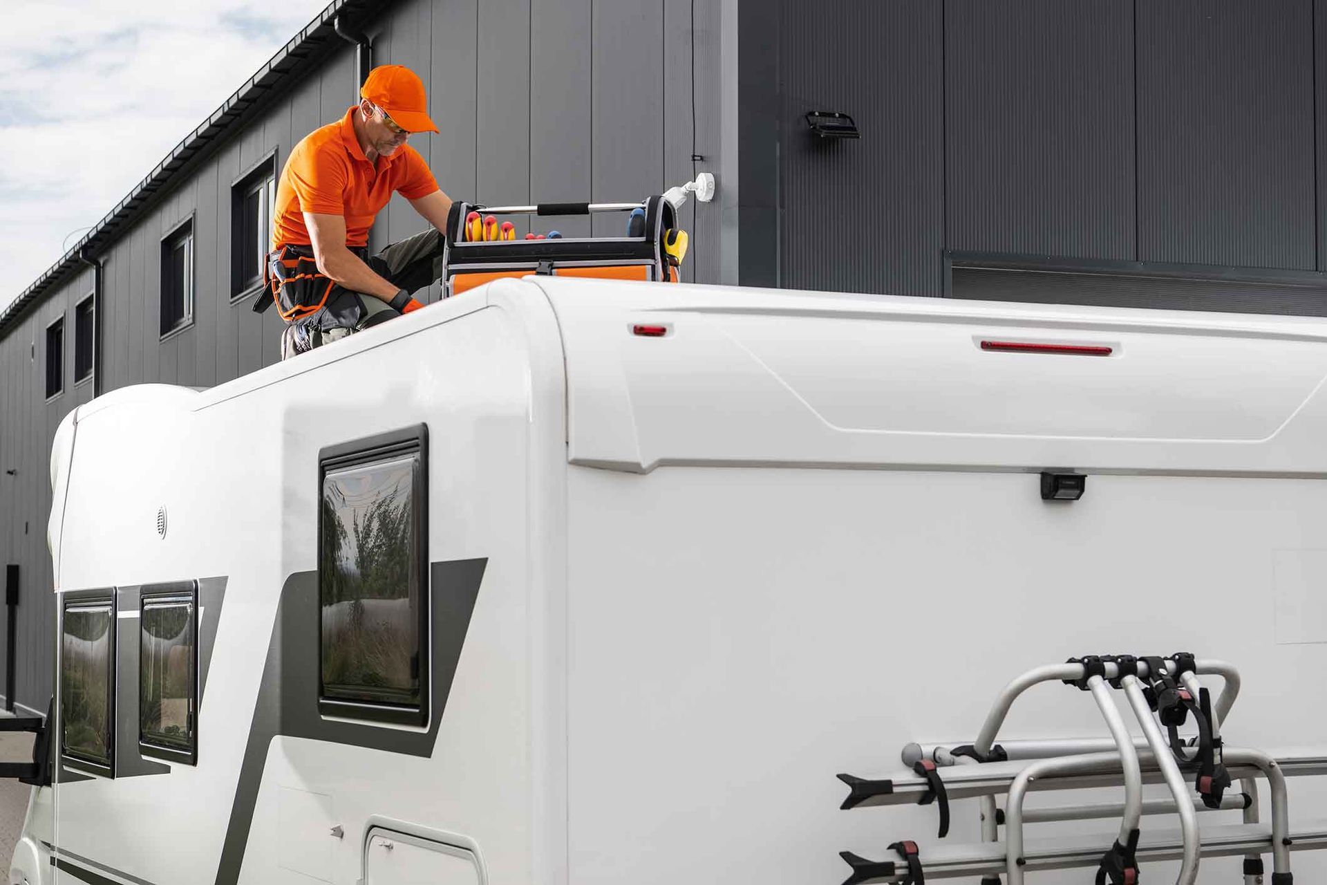 Person in orange shirt working on the roof of a white RV. A building is in the background.