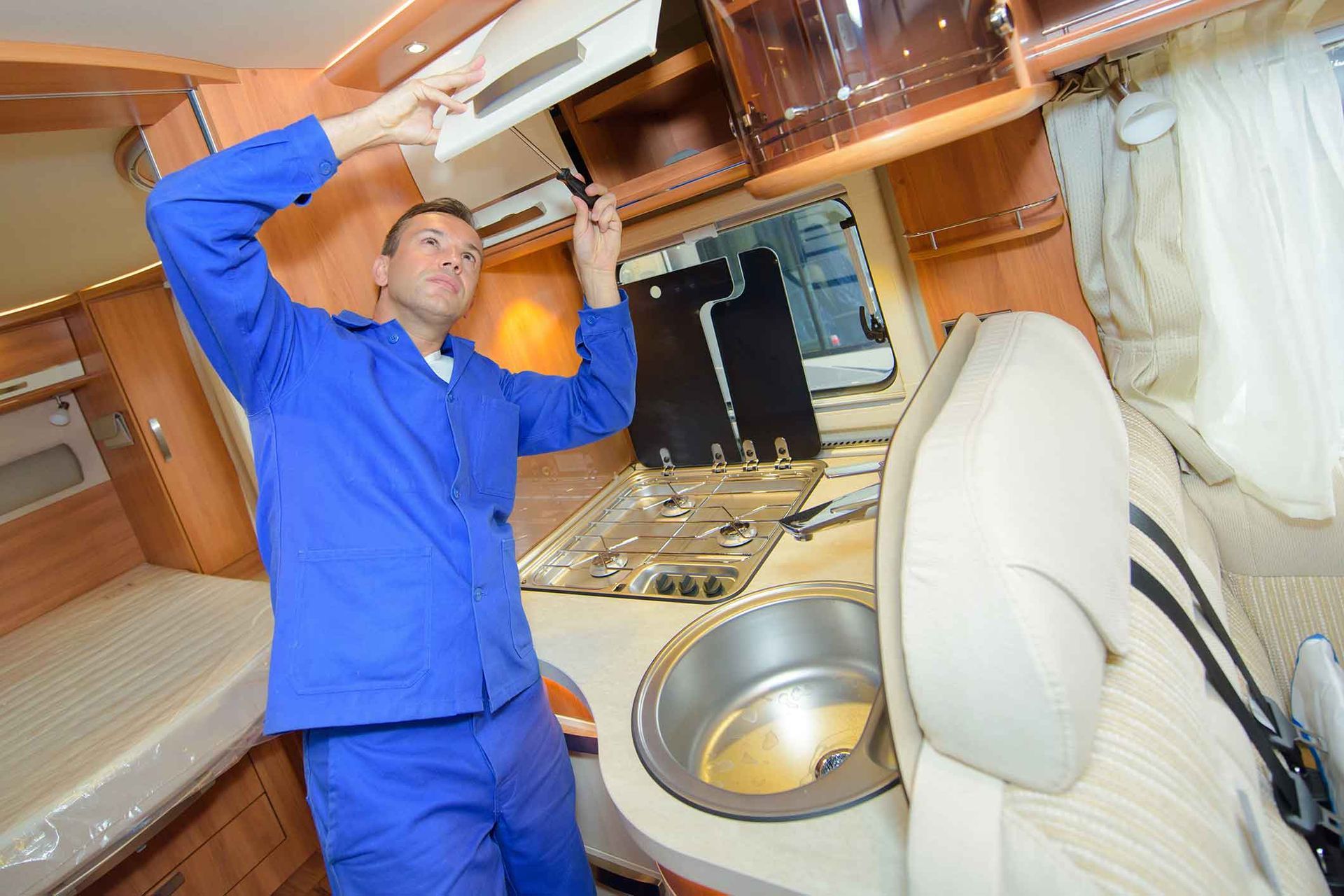 Man in blue coveralls fixing cabinet in RV kitchen.