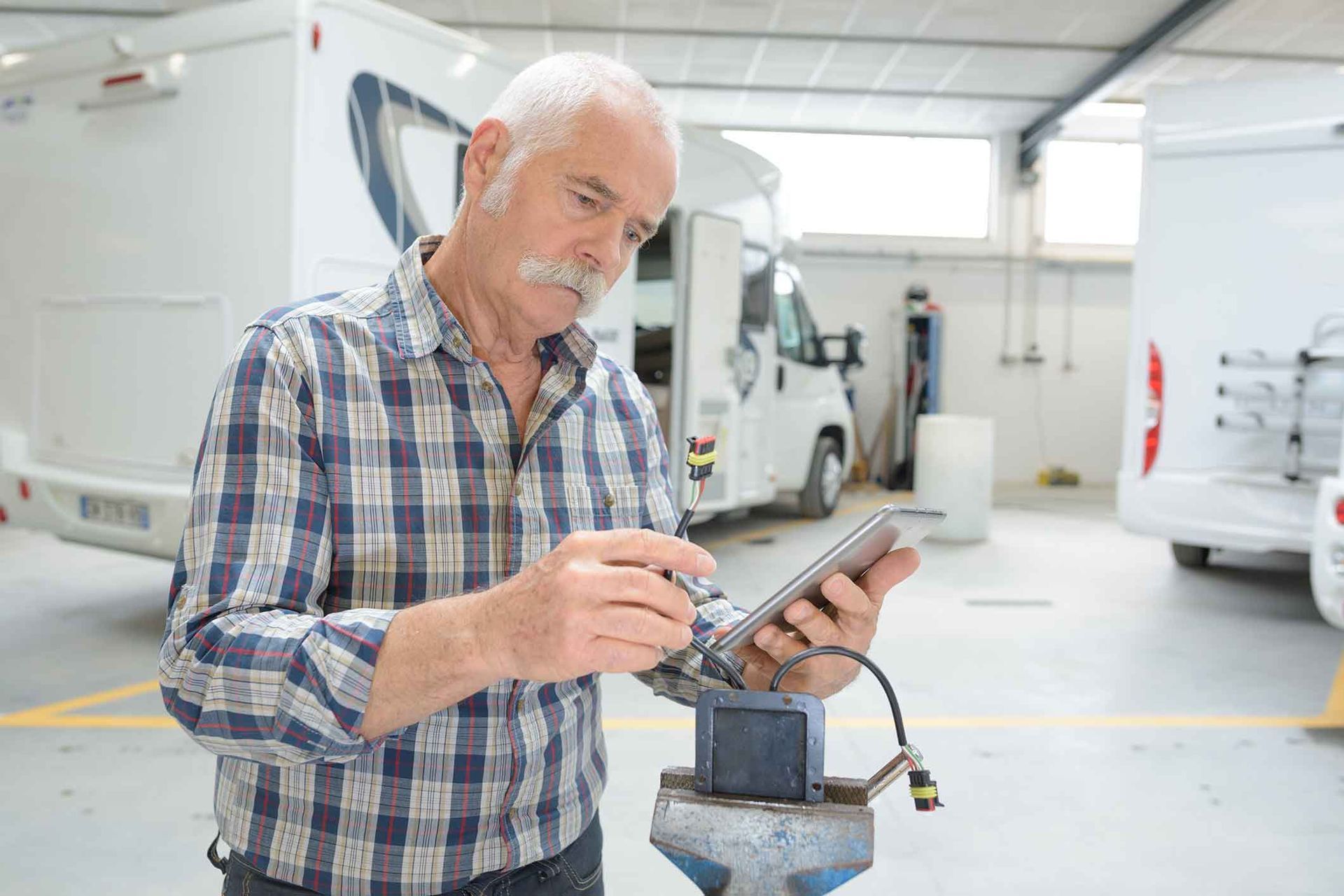 Man in plaid shirt, using tablet while working on a vehicle wire in a workshop, RVs in background.