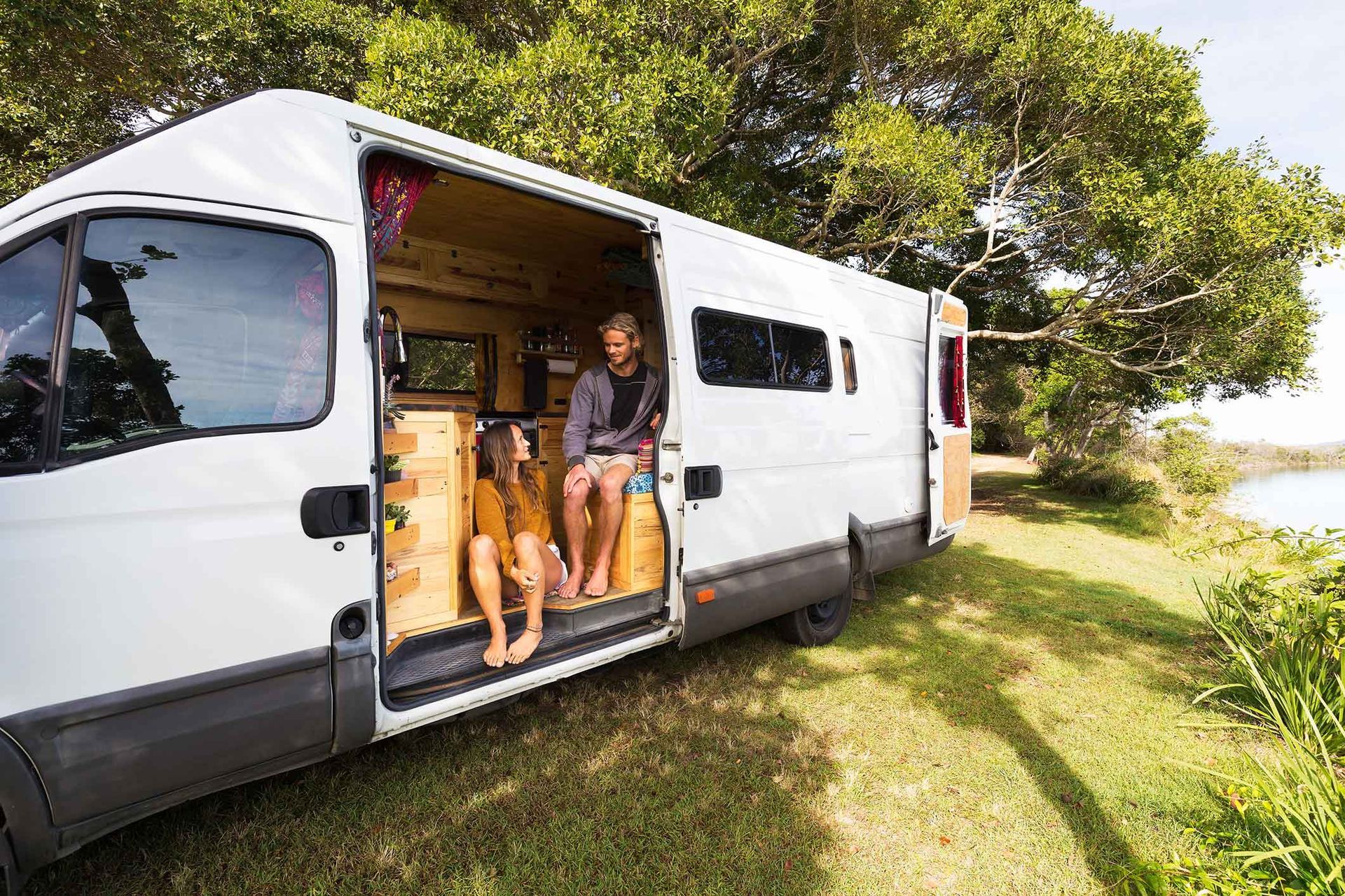 White camper van with open door, two people inside, parked near water and trees.