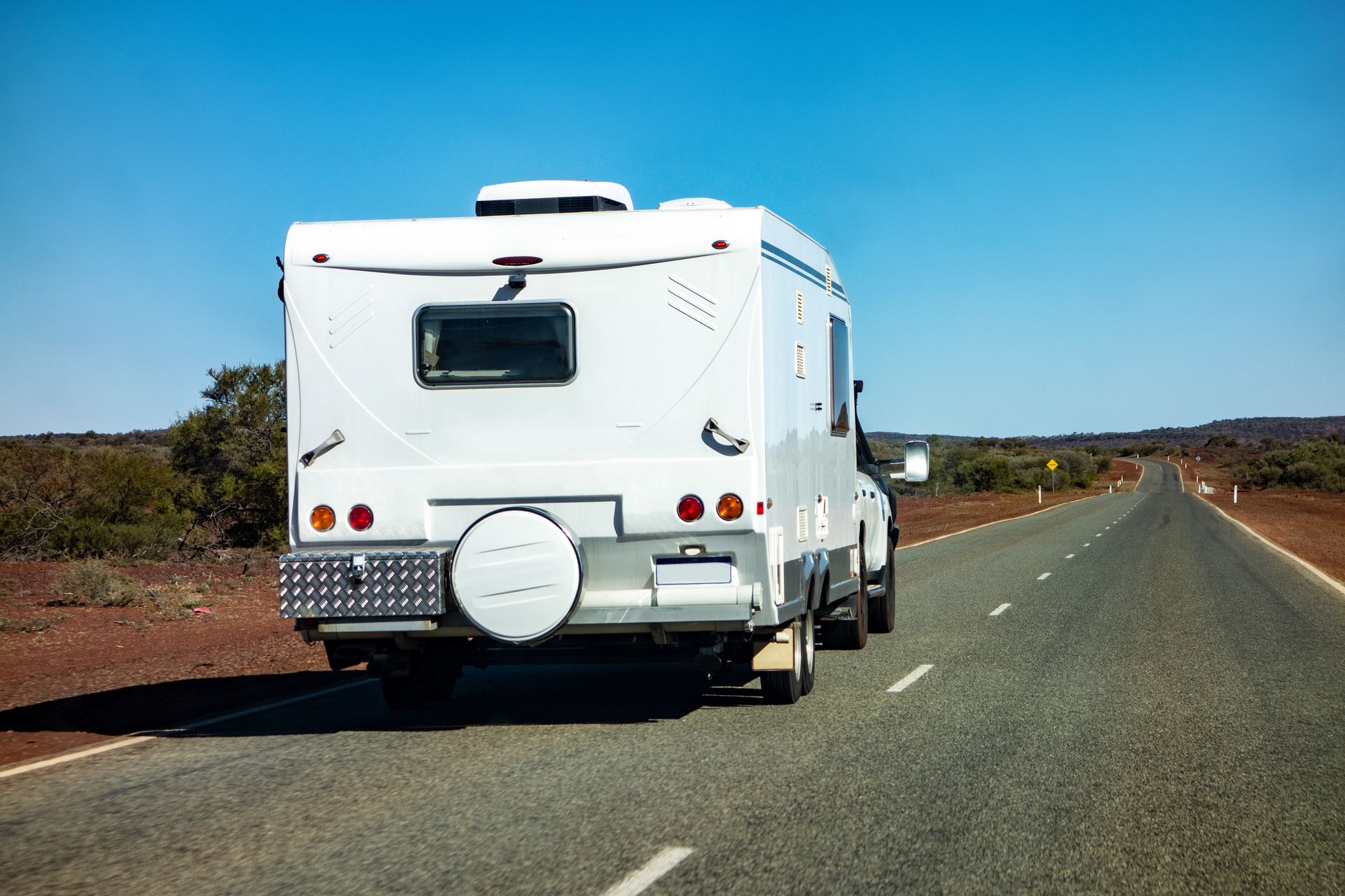 White RV driving on a long, straight road under a clear blue sky.