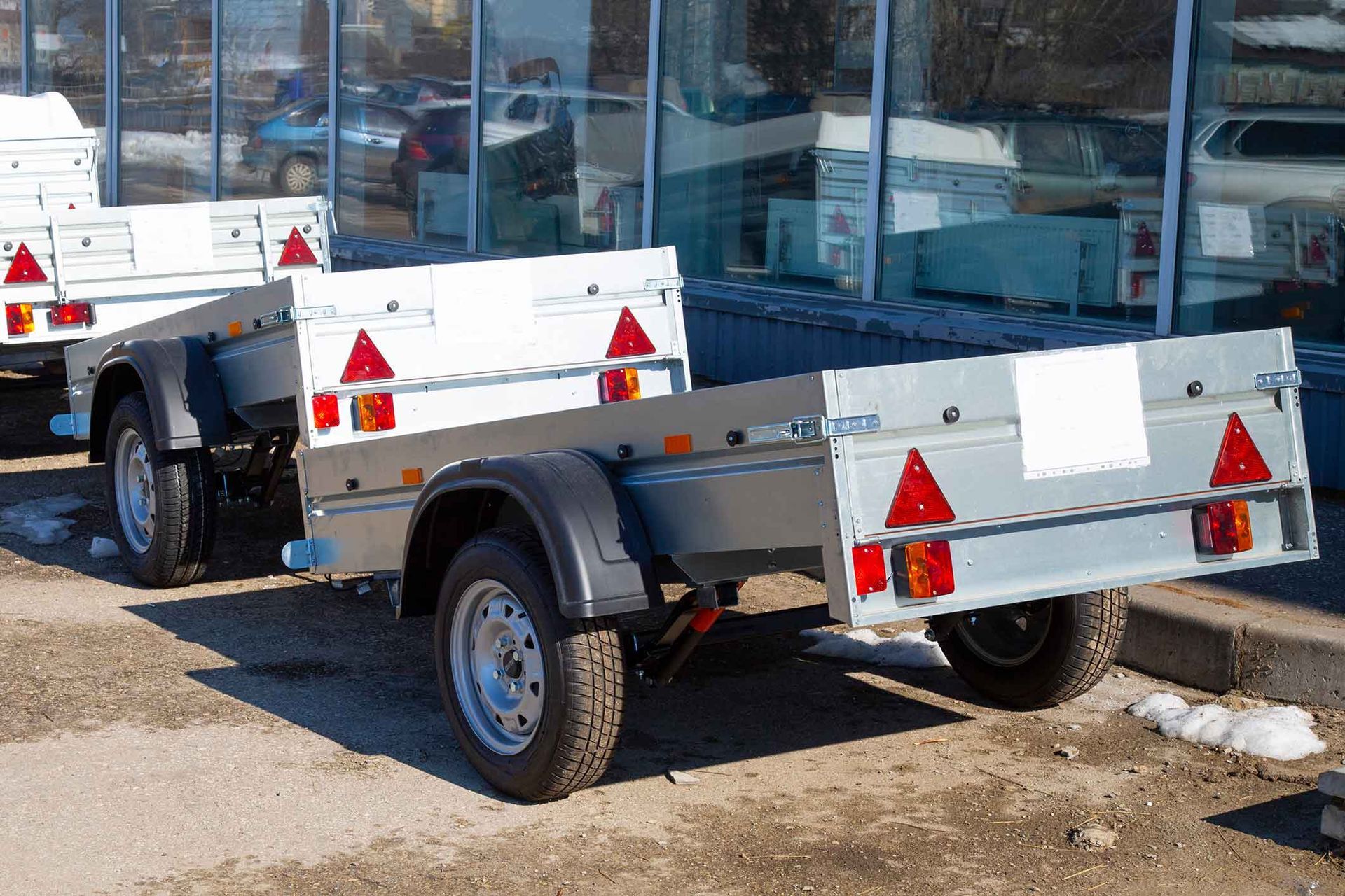 Two silver utility trailers with red triangular reflectors and black tires parked outside a building.