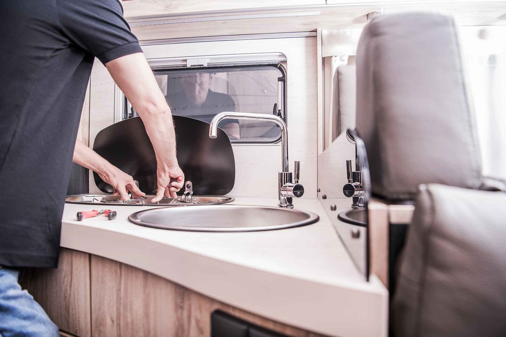 Person fixing a sink in a recreational vehicle. They're using tools, near a window and seating.