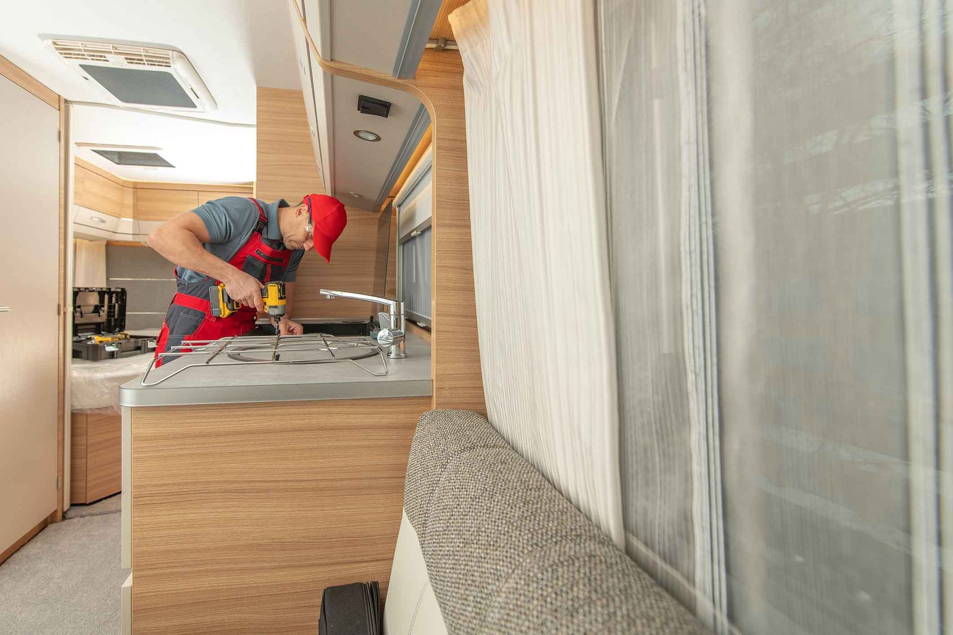 Person in red overalls drilling in a camper van kitchen, next to a window with curtains.