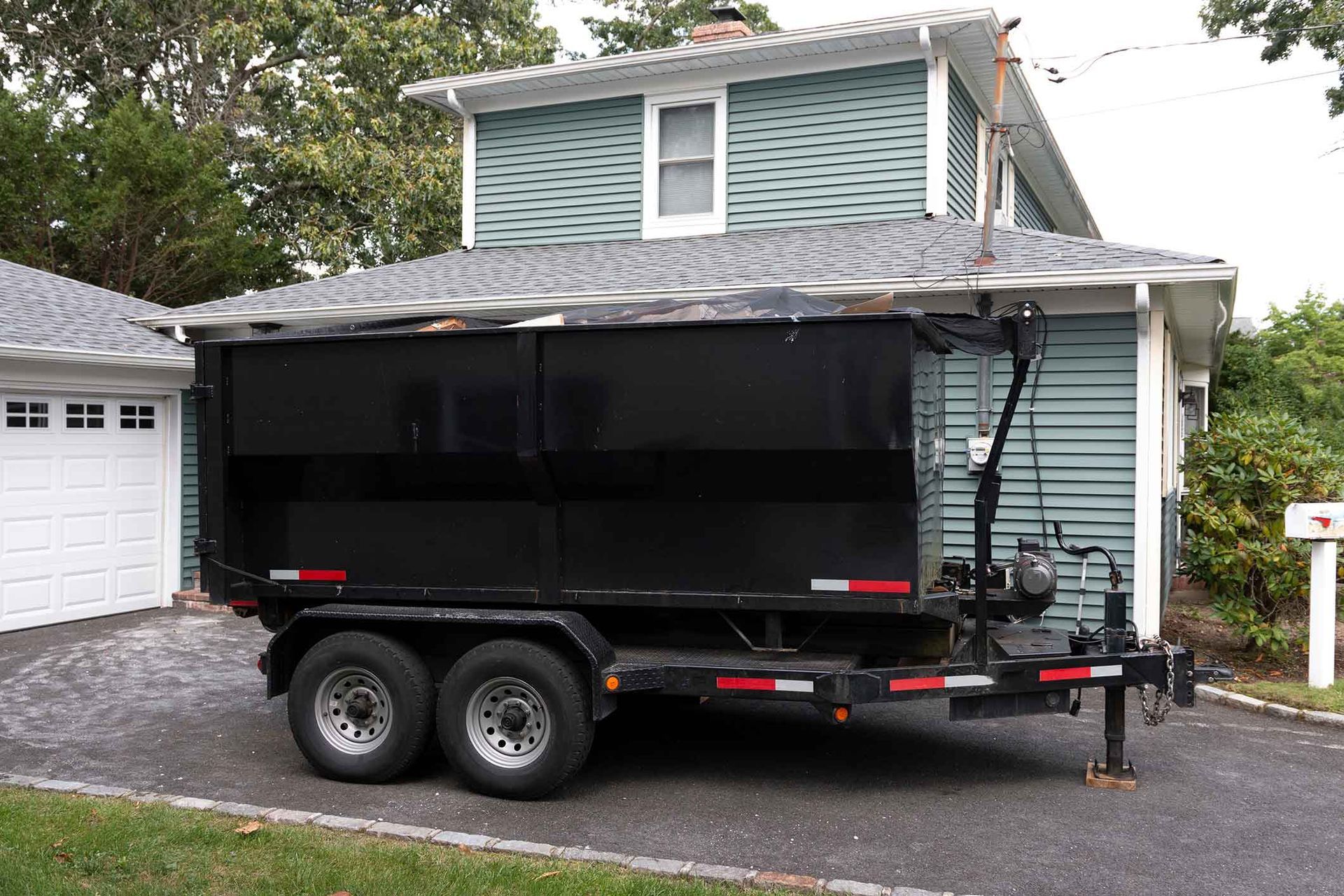 Black dumpster trailer parked next to a house with blue siding and a white garage.