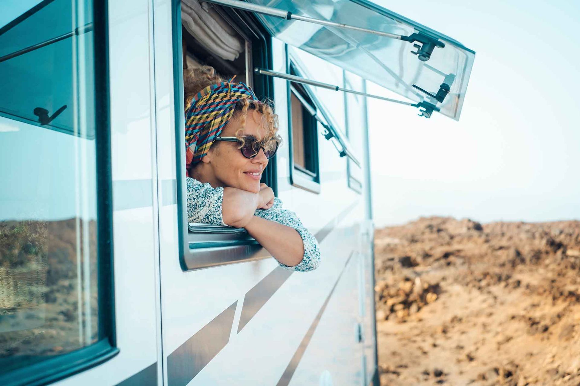 Woman in headscarf and sunglasses leans out of RV window, smiling in a desert setting.