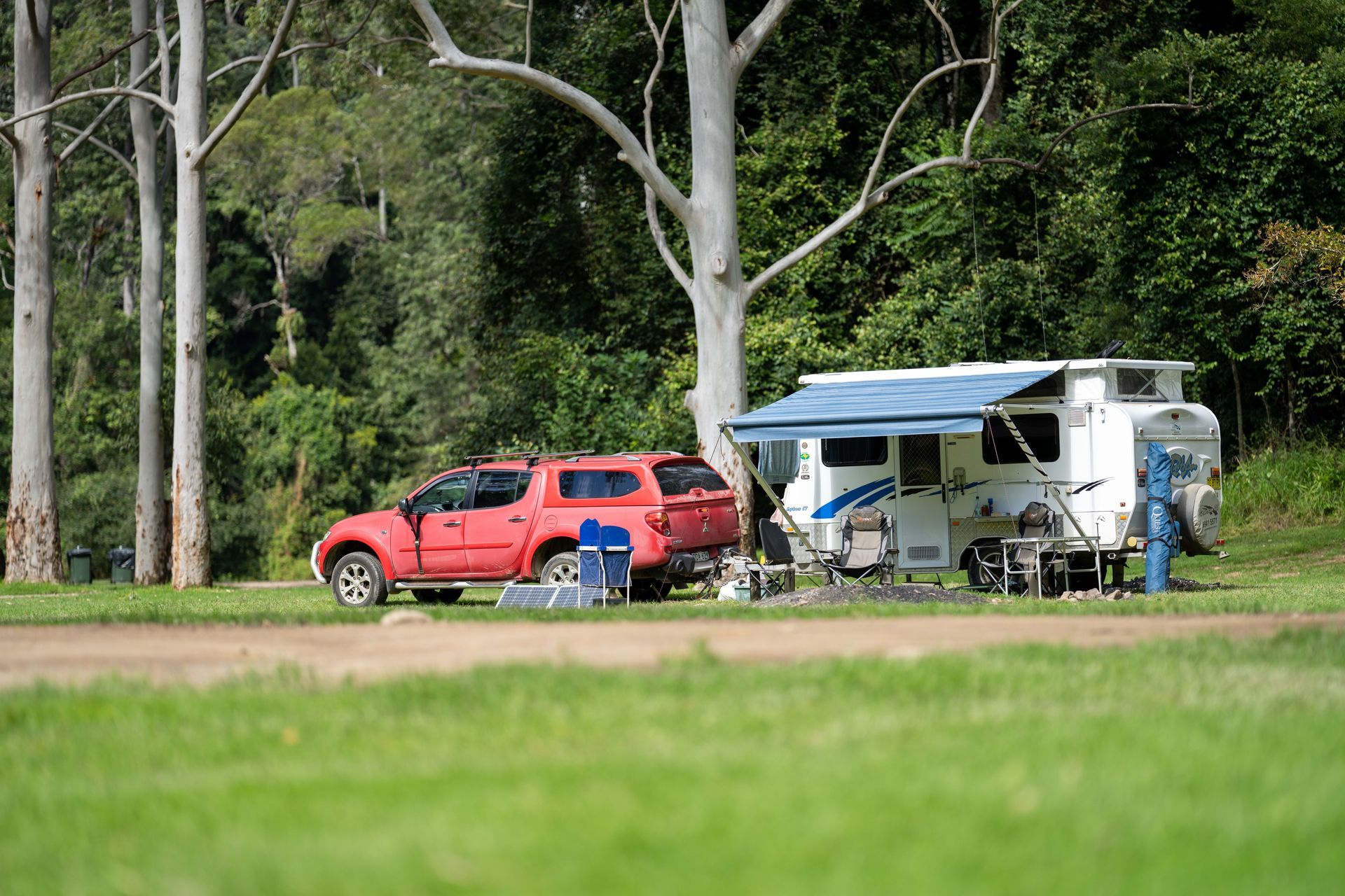 Red truck and camper trailer parked on grass in a campground, trees in the background.
