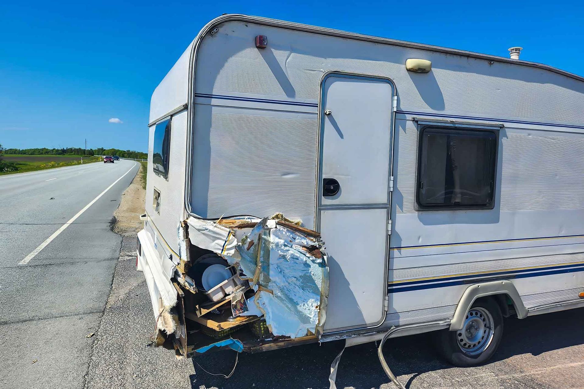 Damaged white travel trailer parked on the side of a road after an accident; blue sky.