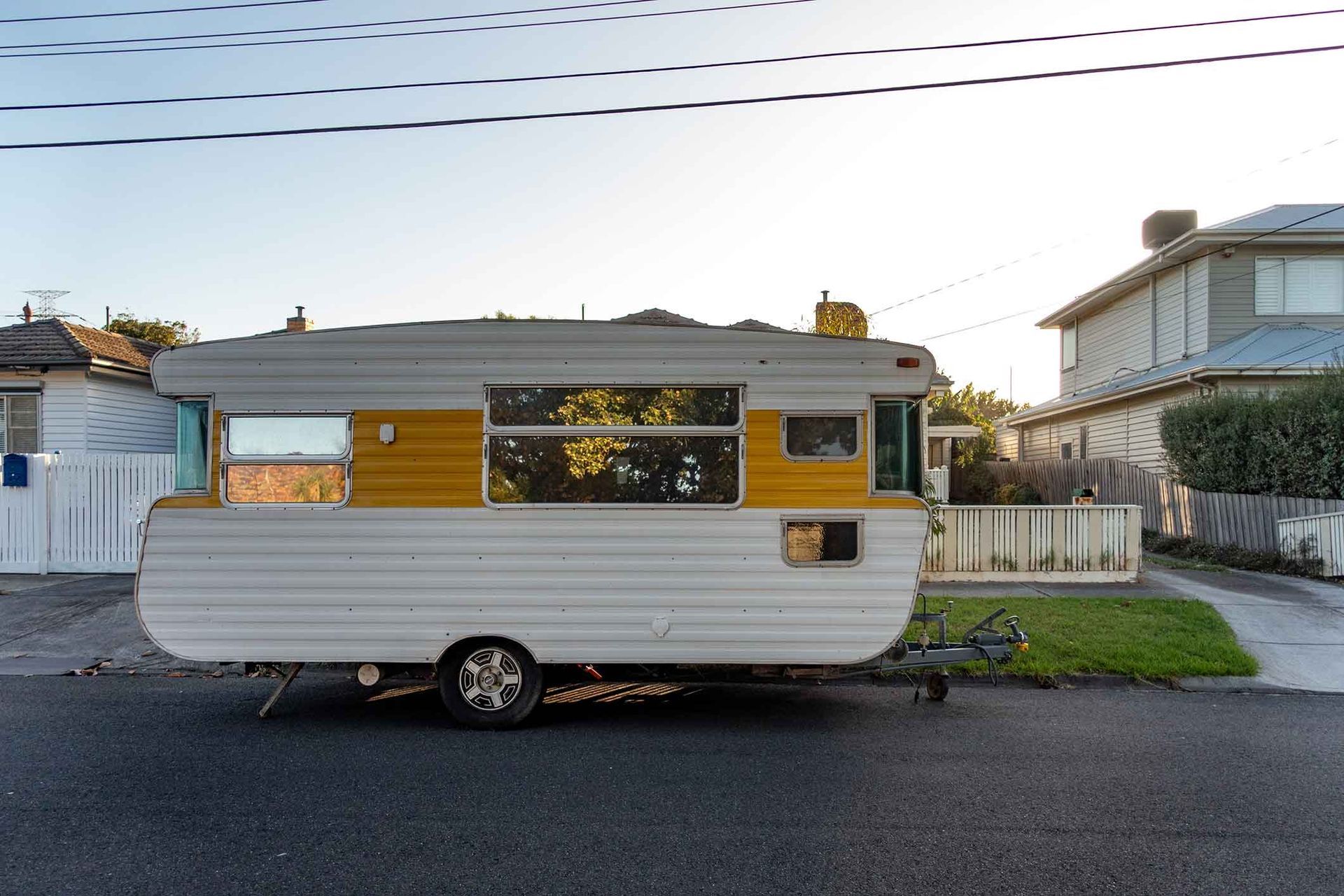 White and yellow camper trailer parked on a street in front of houses; power lines above.