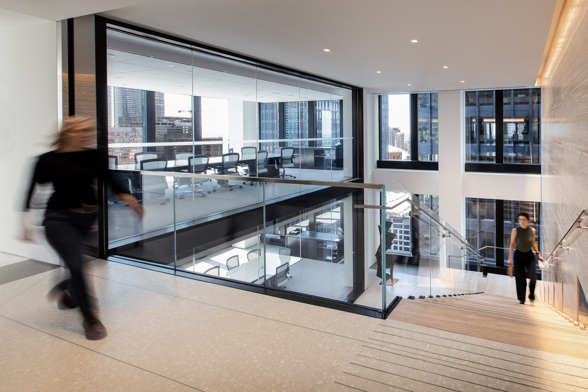 Modern office stairwell with glass railings, large windows, and people walking on a bright mezzanine.