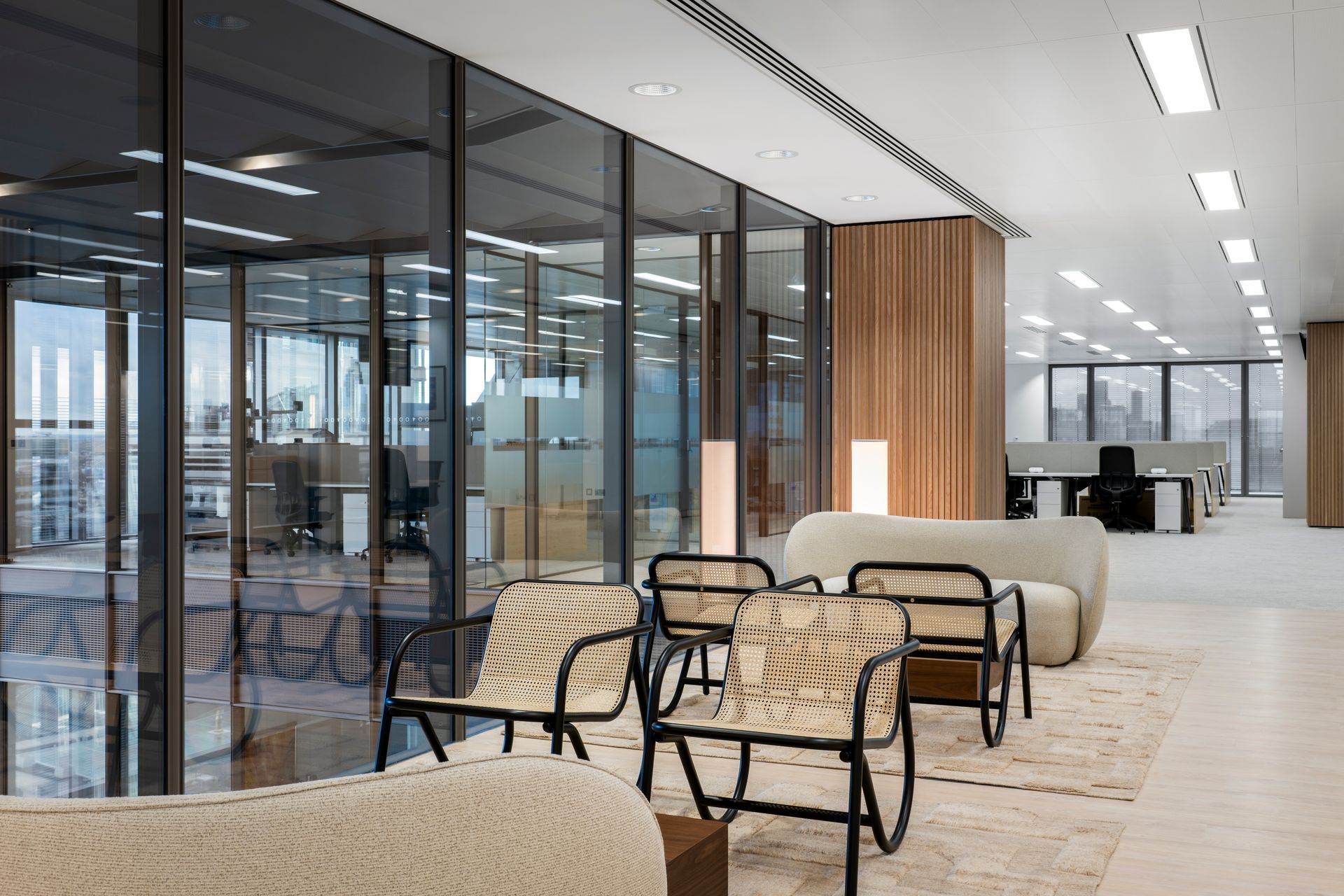 Modern office lounge with glass walls, woven chairs, beige sofas, and bright open workspace