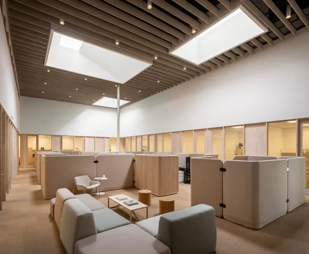 Modern office lounge with beige seating, wood partitions, and skylights in a bright open workspace