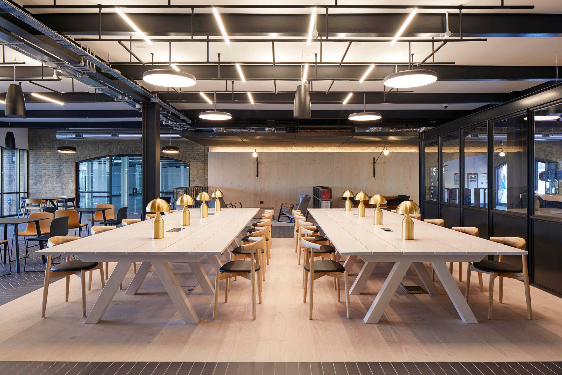 Modern office break room with long white tables, benches, stools, and hanging lights