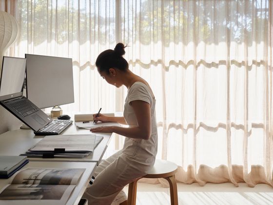 Person writing at a desk beside a laptop in a bright room with sheer curtains.