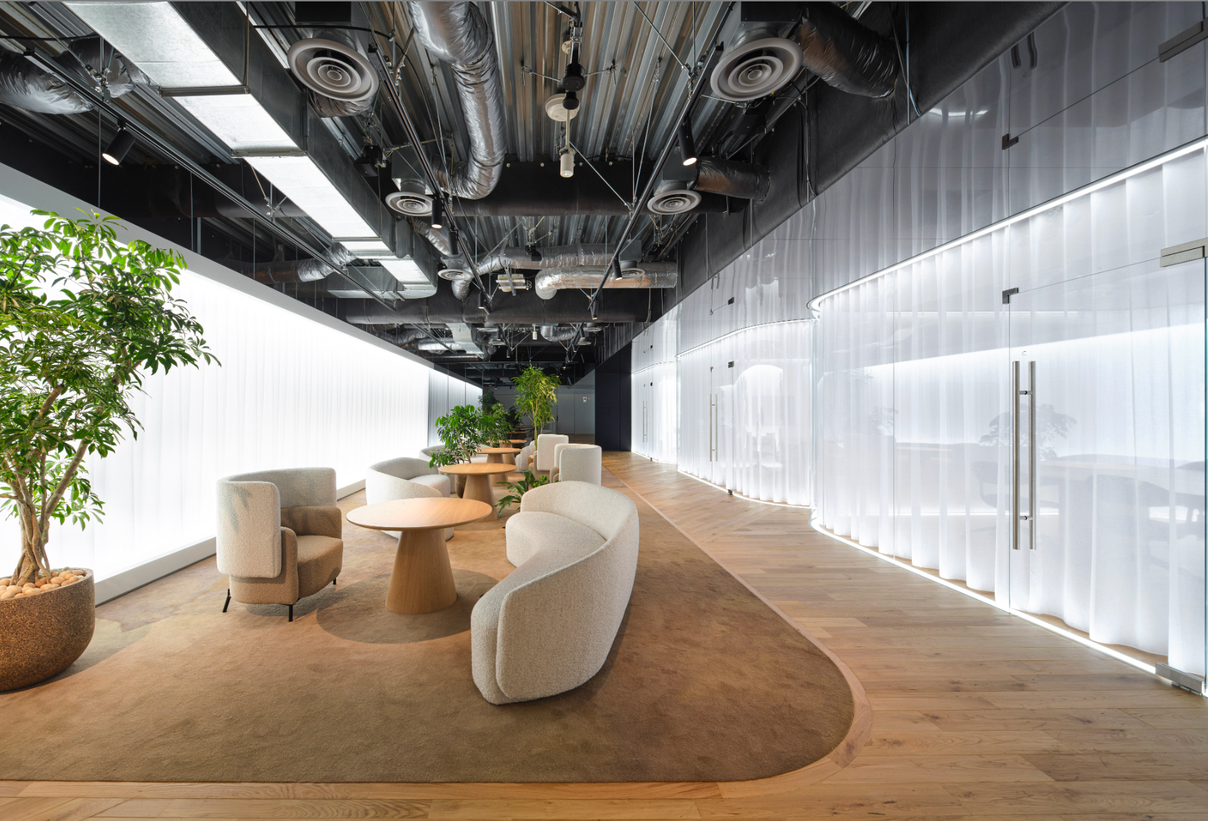 Modern office lounge with curved seating, a round table, wood flooring, and exposed ceiling ducts