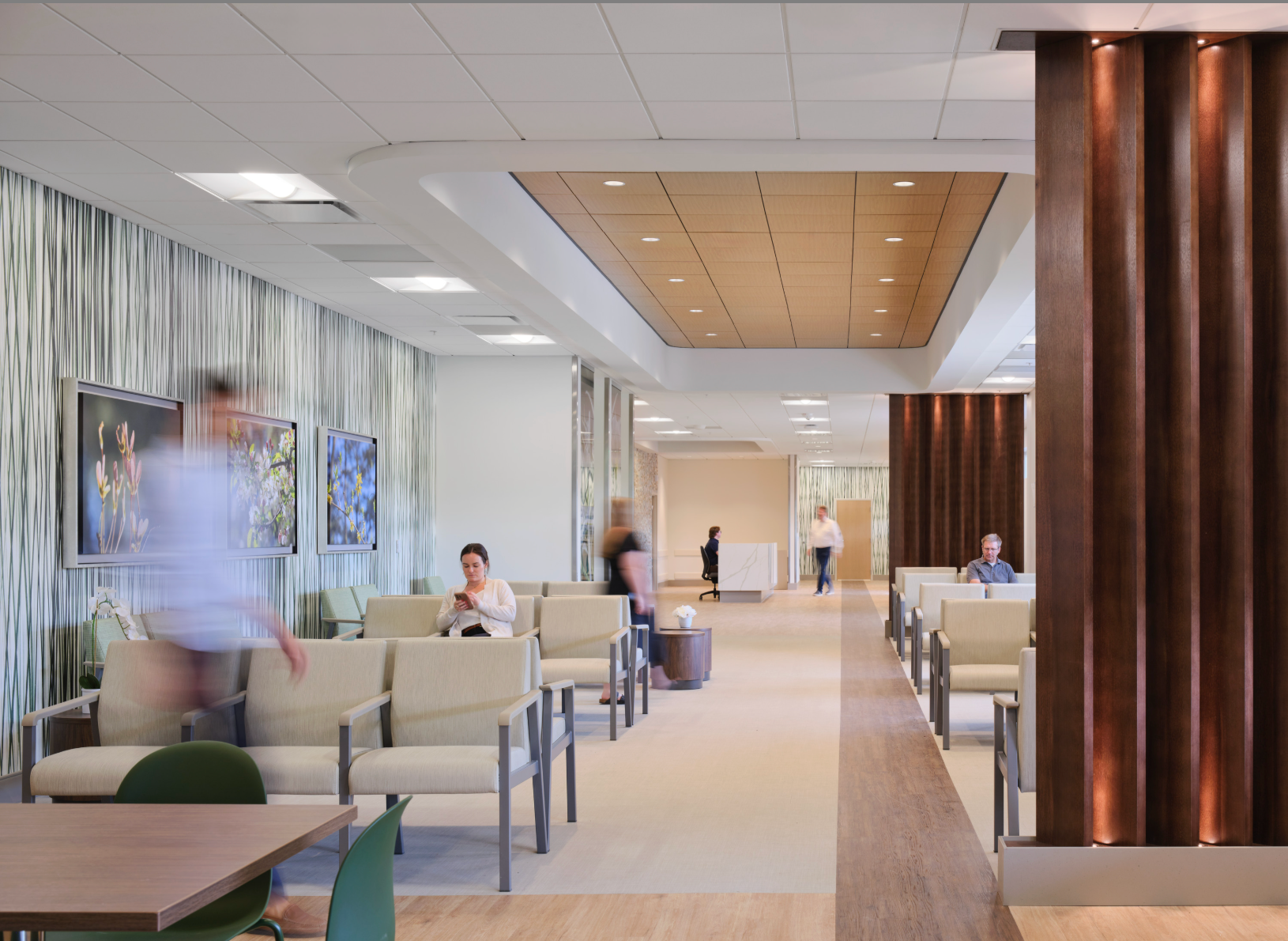 Modern hospital waiting room with beige chairs, wood accents, and a few seated people