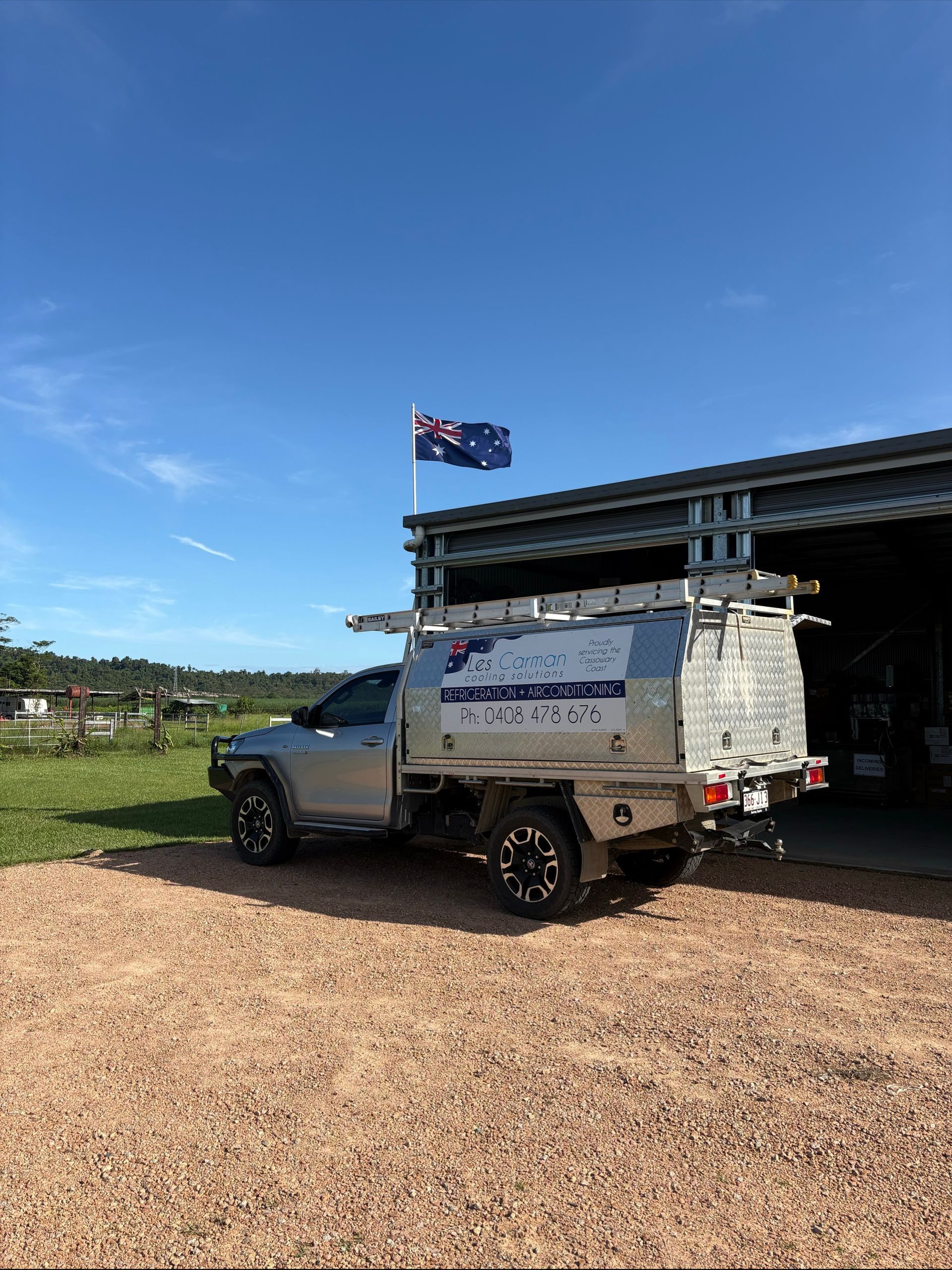 Silver Les Carman truck with tool storage, Australian flag, parked in front of a shed on a gravel surface under a blue sky.