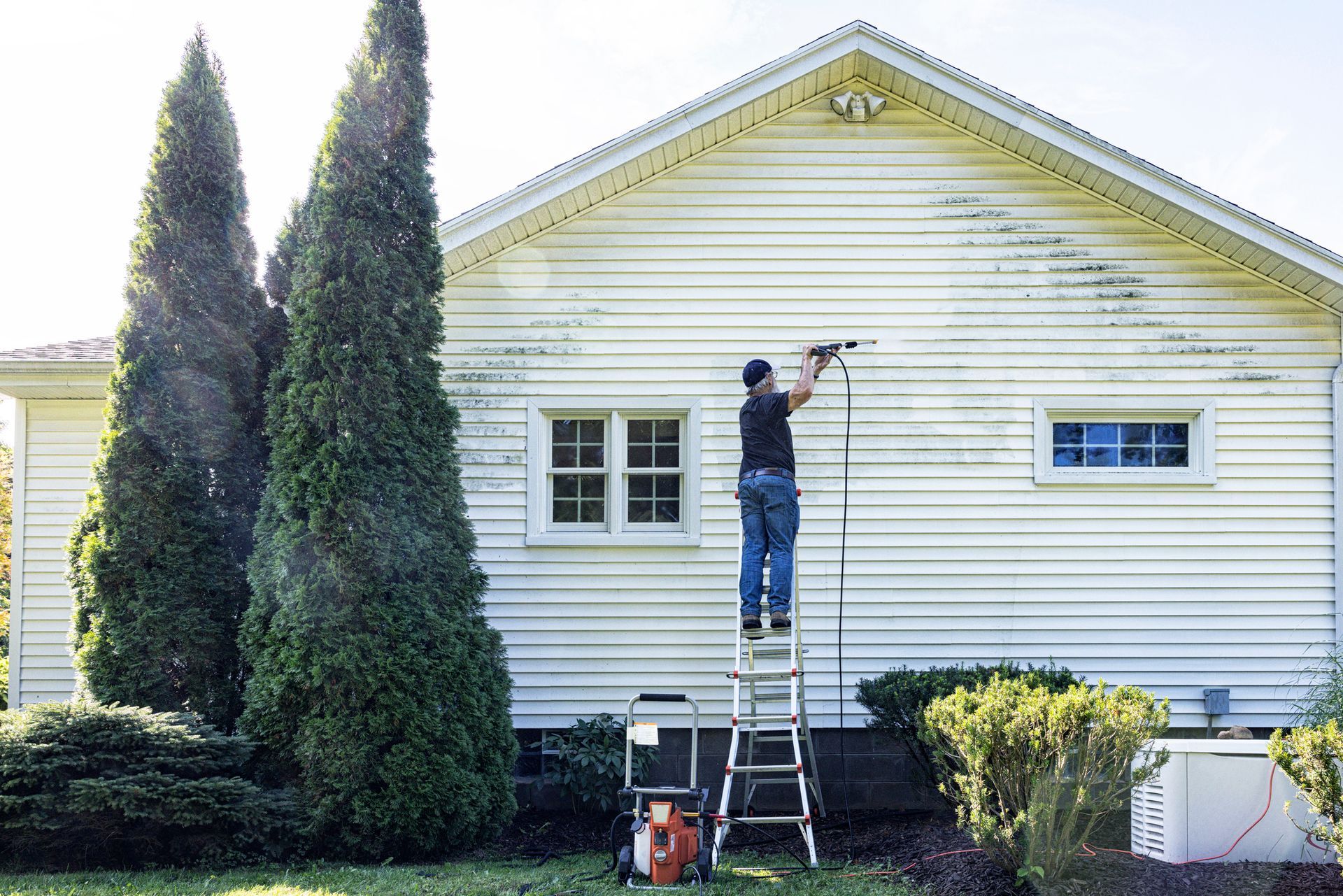 A man is standing on a ladder pressure washing the siding from the house wall.