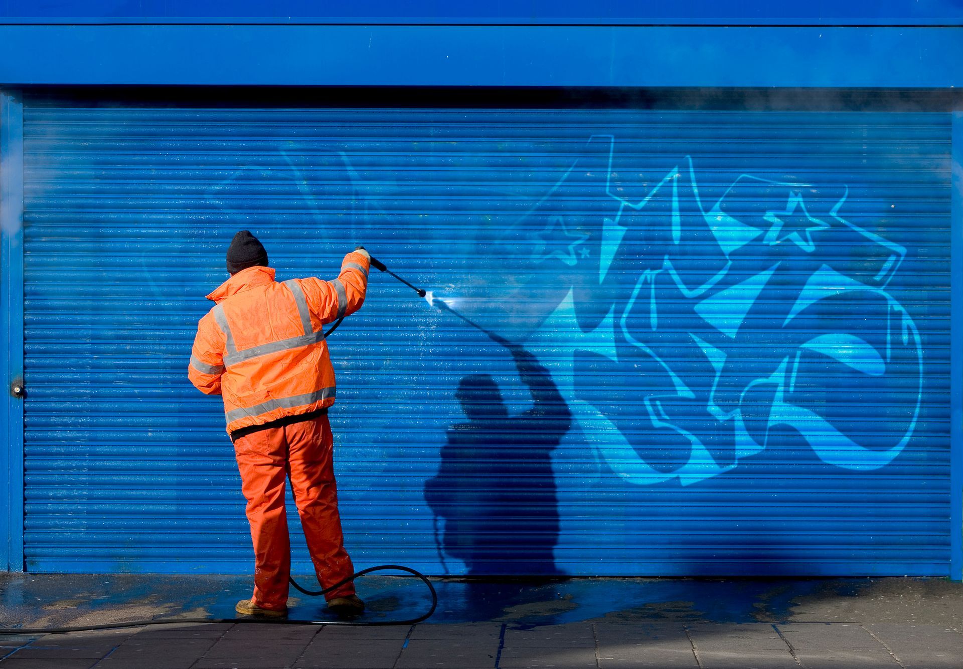 A man is cleaning graffiti on a blue garage door with a high pressure washer.