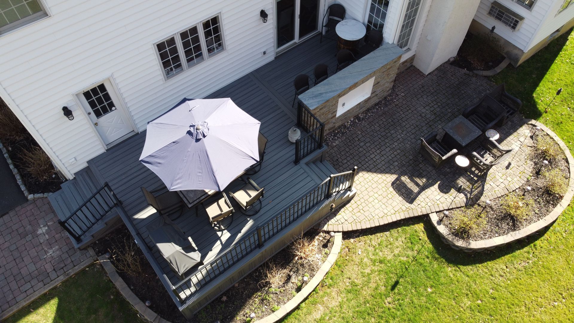 Aerial view of a backyard with a hot tub, deck, seating, and umbrella, next to a house with a gray roof.