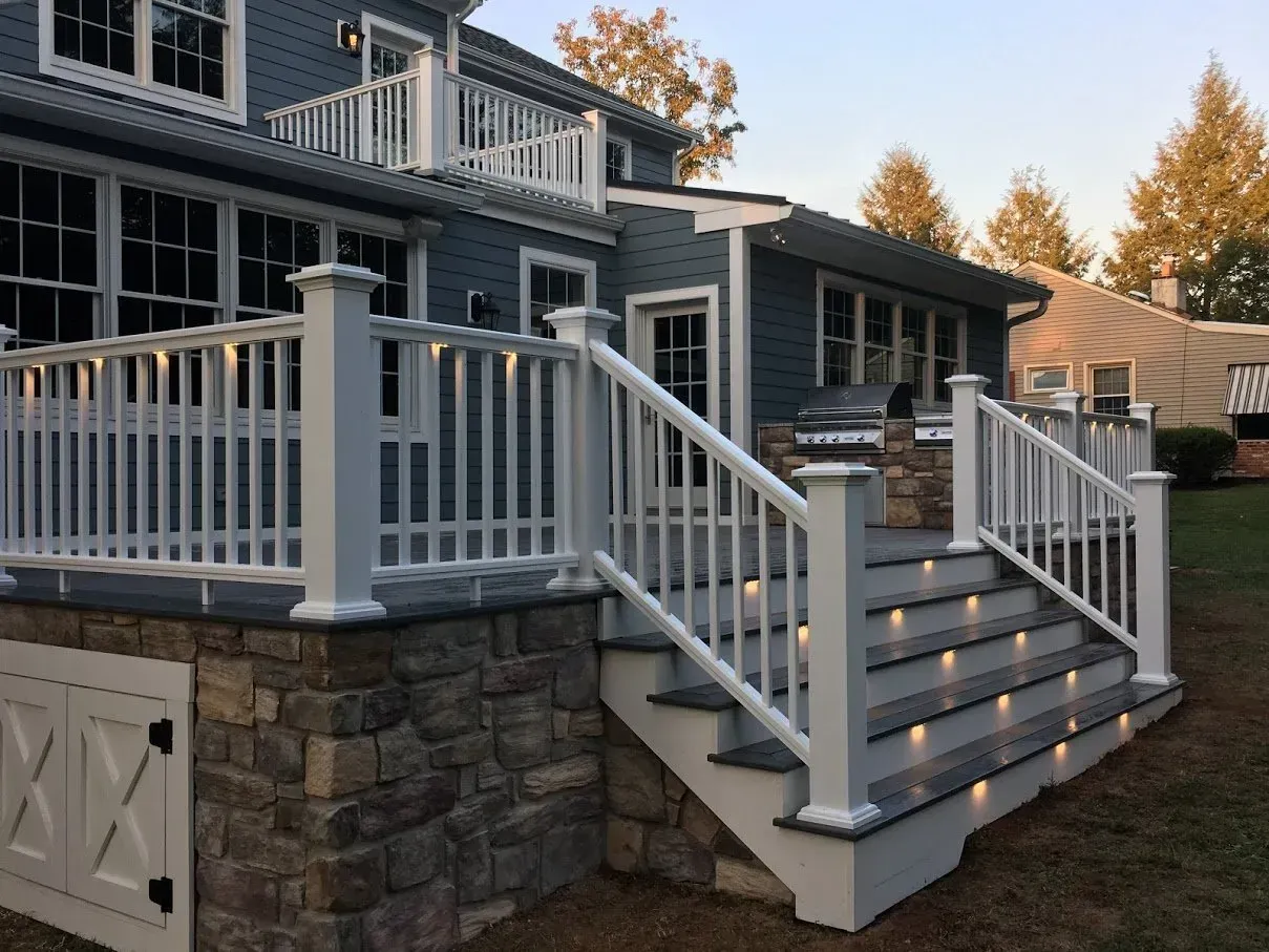 Backyard deck with gray steps, white railings, and stone facade, with a grill and built-in lights.