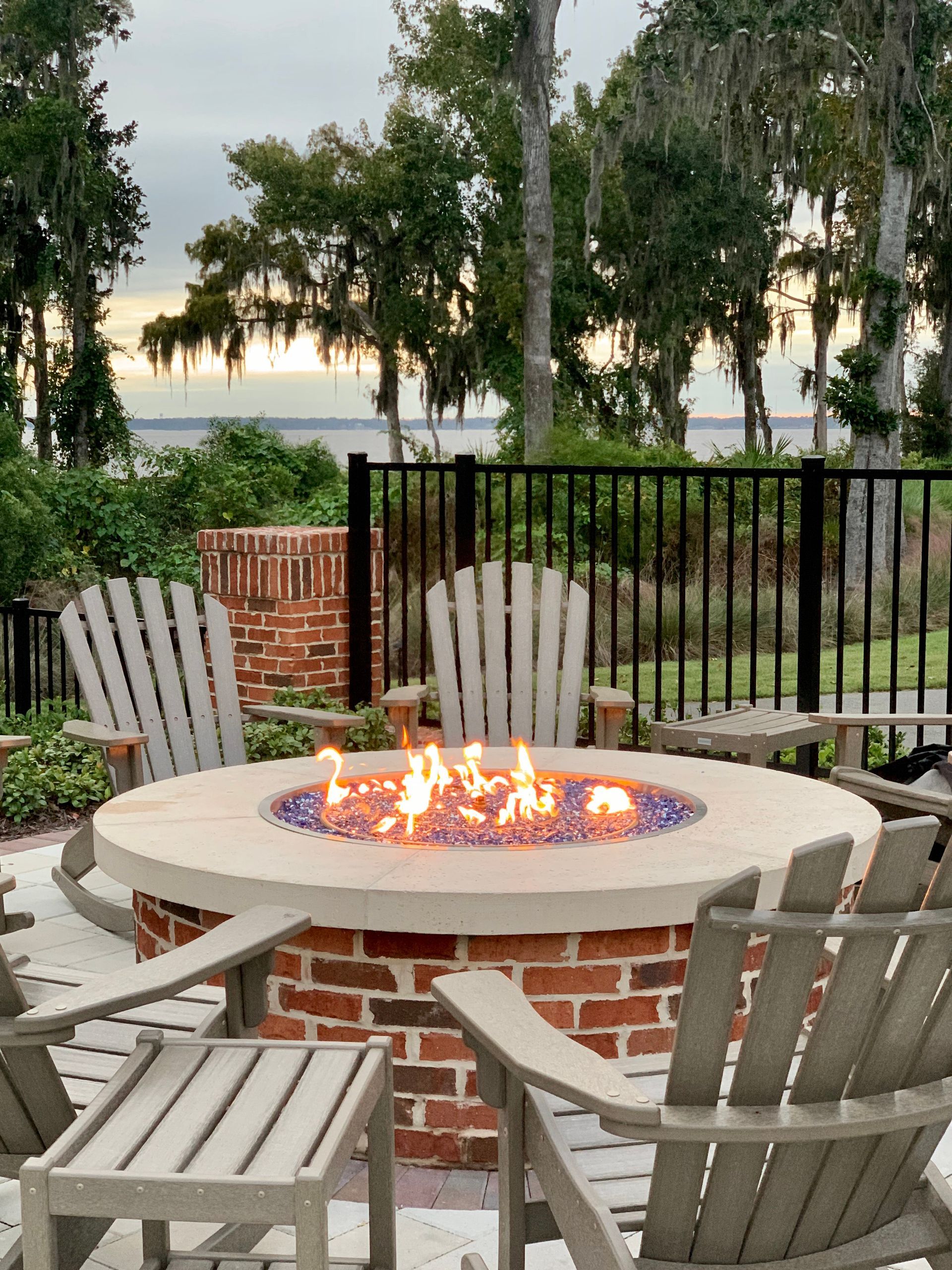 Patio fire pit with wooden chairs, brick base, and lakefront trees at dusk